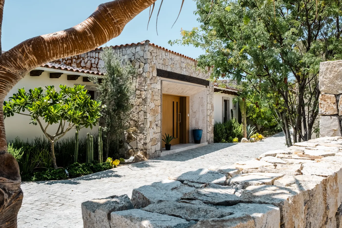 Stone-clad entrance with a wood pivot door and cobblestone path through a desert garden, Casa Querencia 26 in Los Cabos
