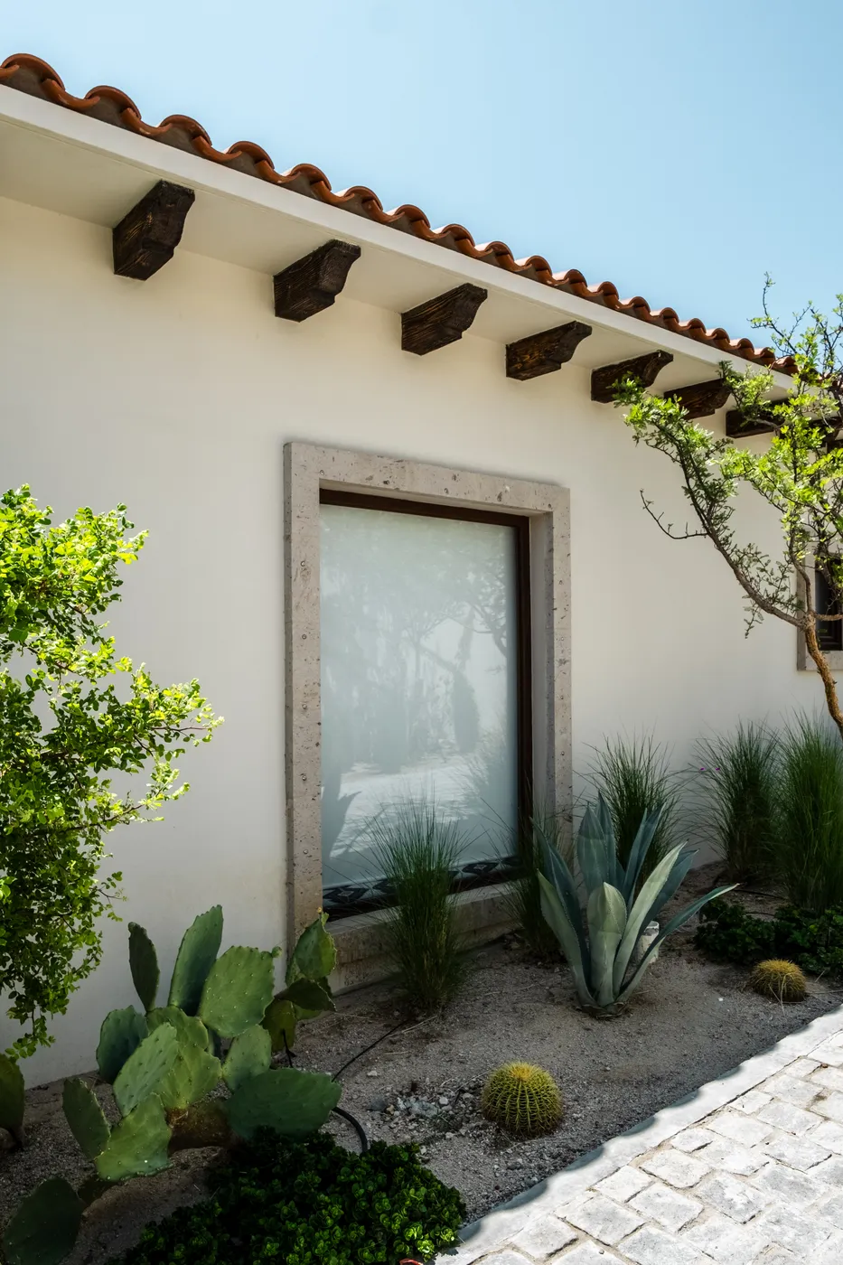 Stone-framed window in a white stucco wall under a clay-tile eave with wood corbels, agaves and cacti below