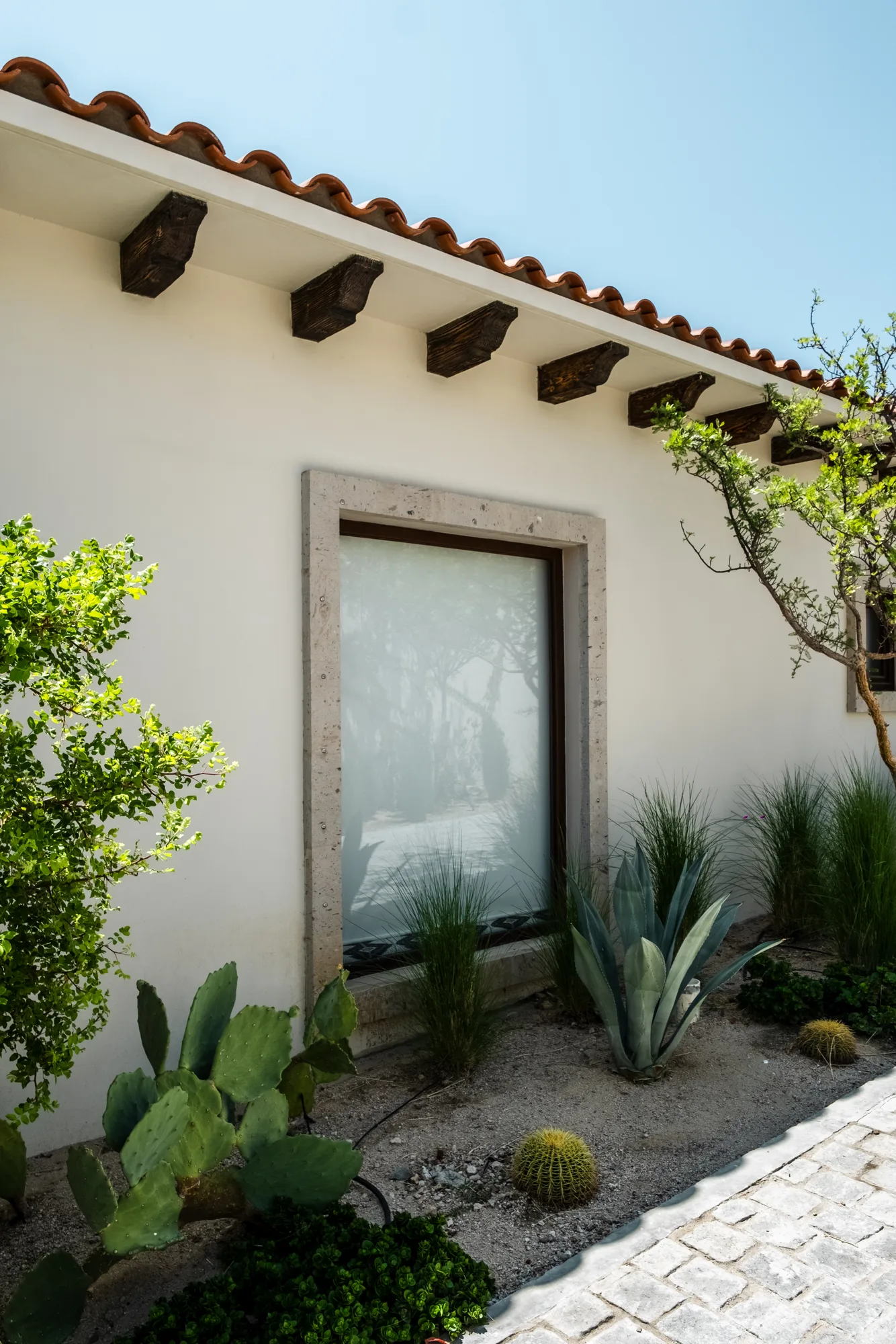 Stone-framed window in a white stucco wall under a clay-tile eave with wood corbels, agaves and cacti below