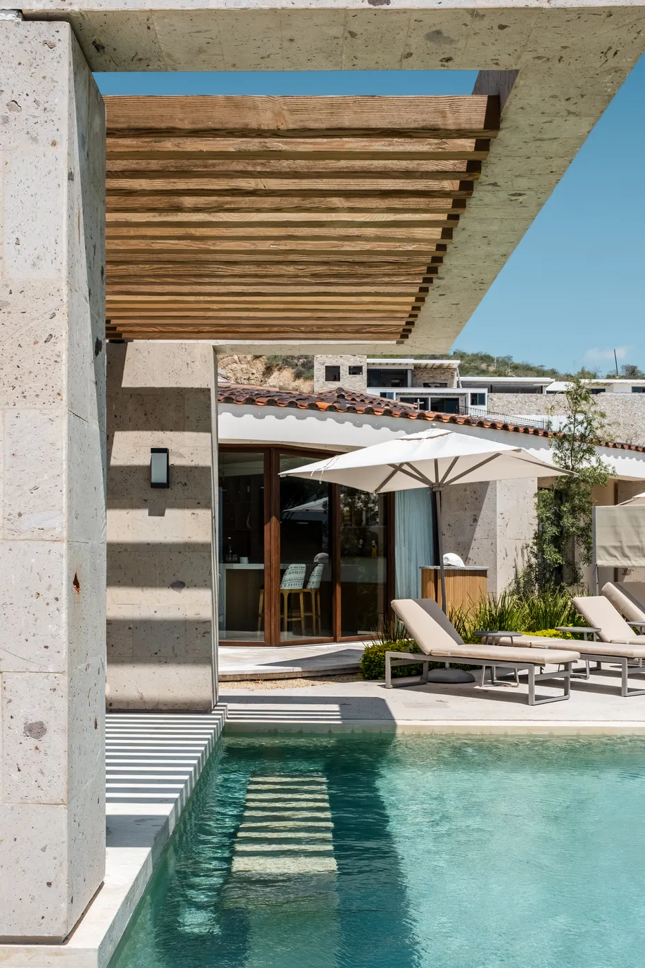View through a wood-slat pergola on stone columns toward the pool and a shaded terrace, hillside villas beyond