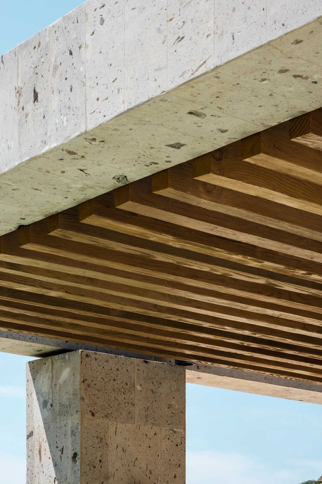 Detail of a cantilevered pergola, a stone beam over timber slats on a stone column against a clear sky