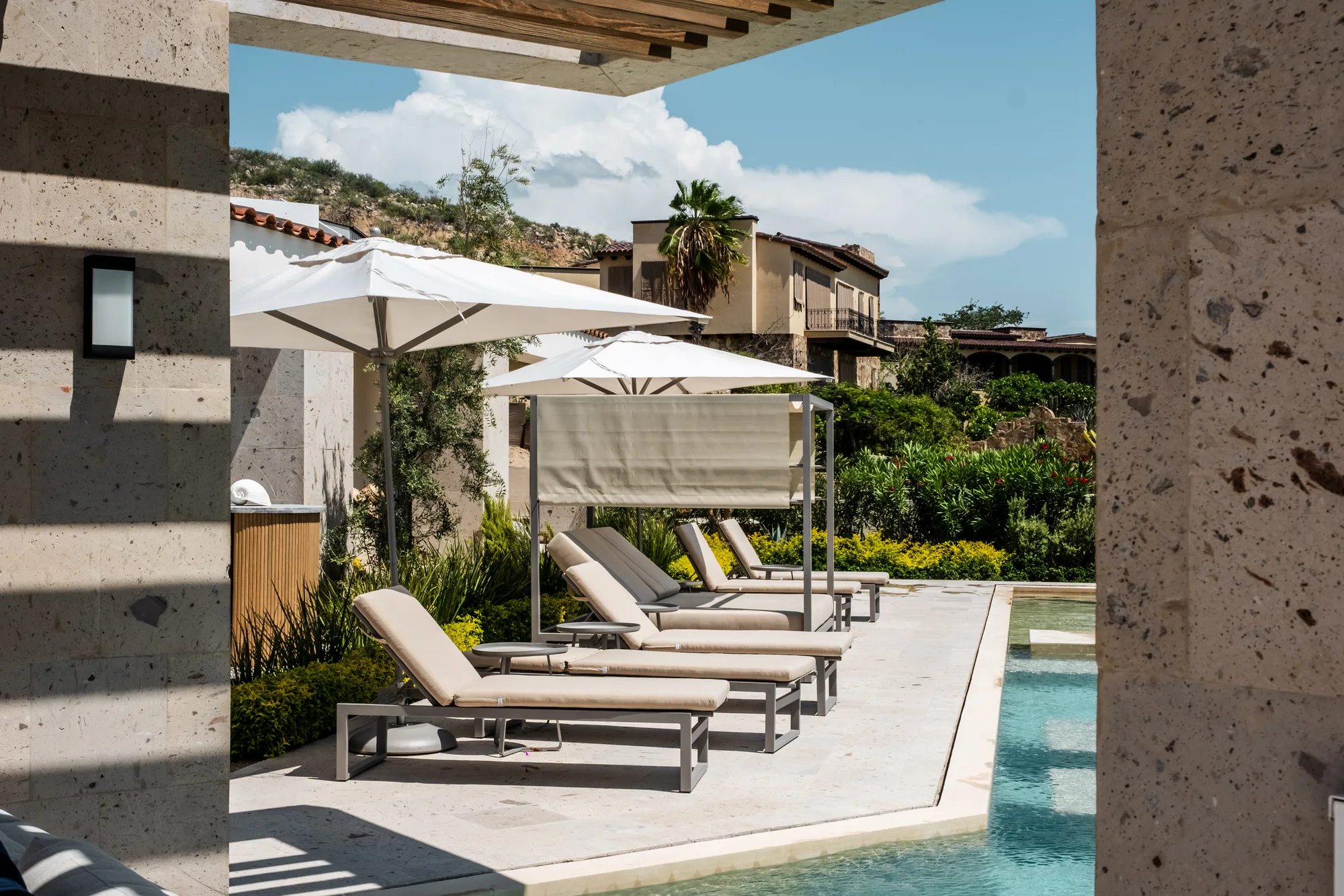 Poolside loungers and white umbrellas framed between travertine columns, hillside villas and a palm beyond