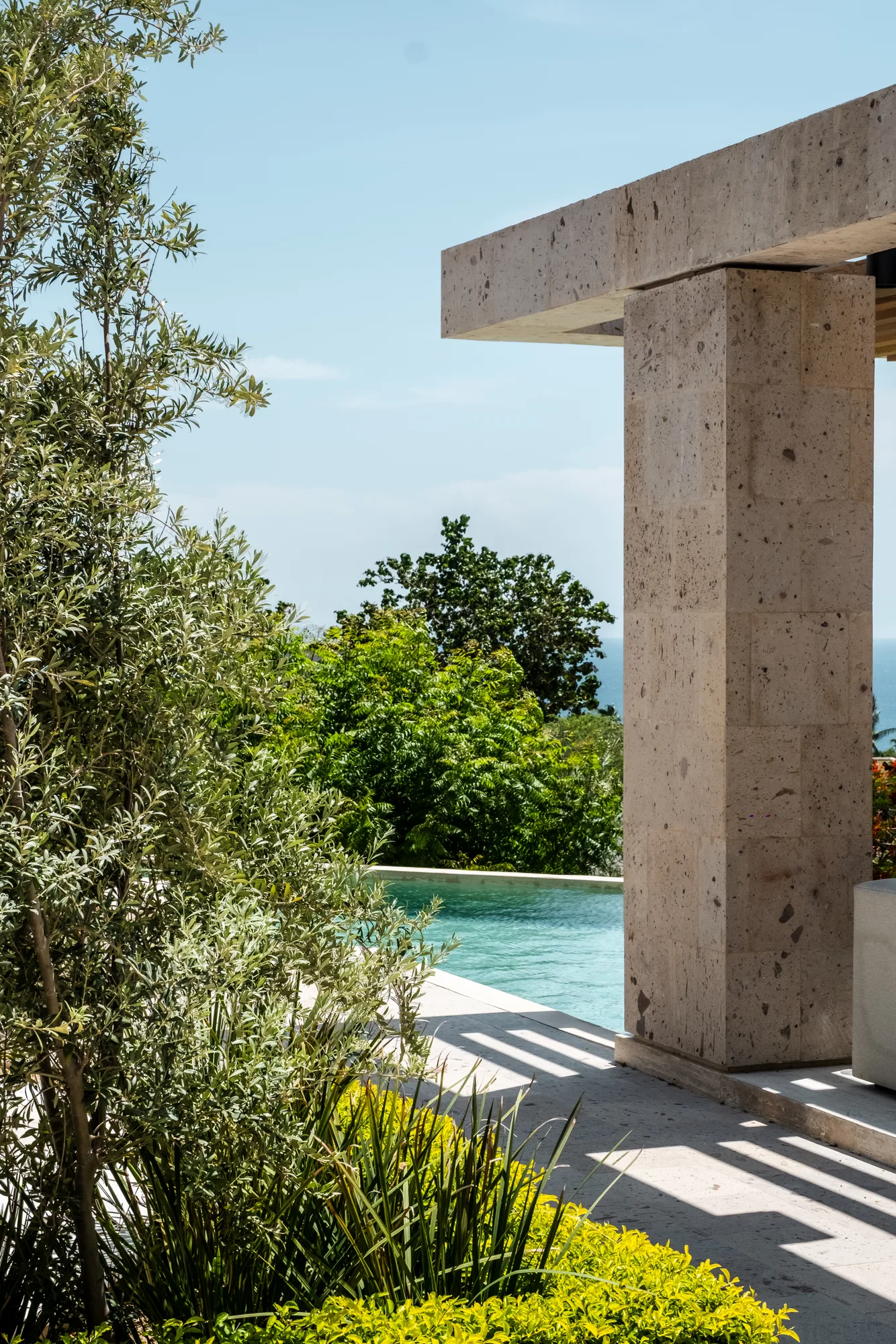 Travertine post and beam framing the infinity pool edge and the sea horizon, an olive tree in the foreground