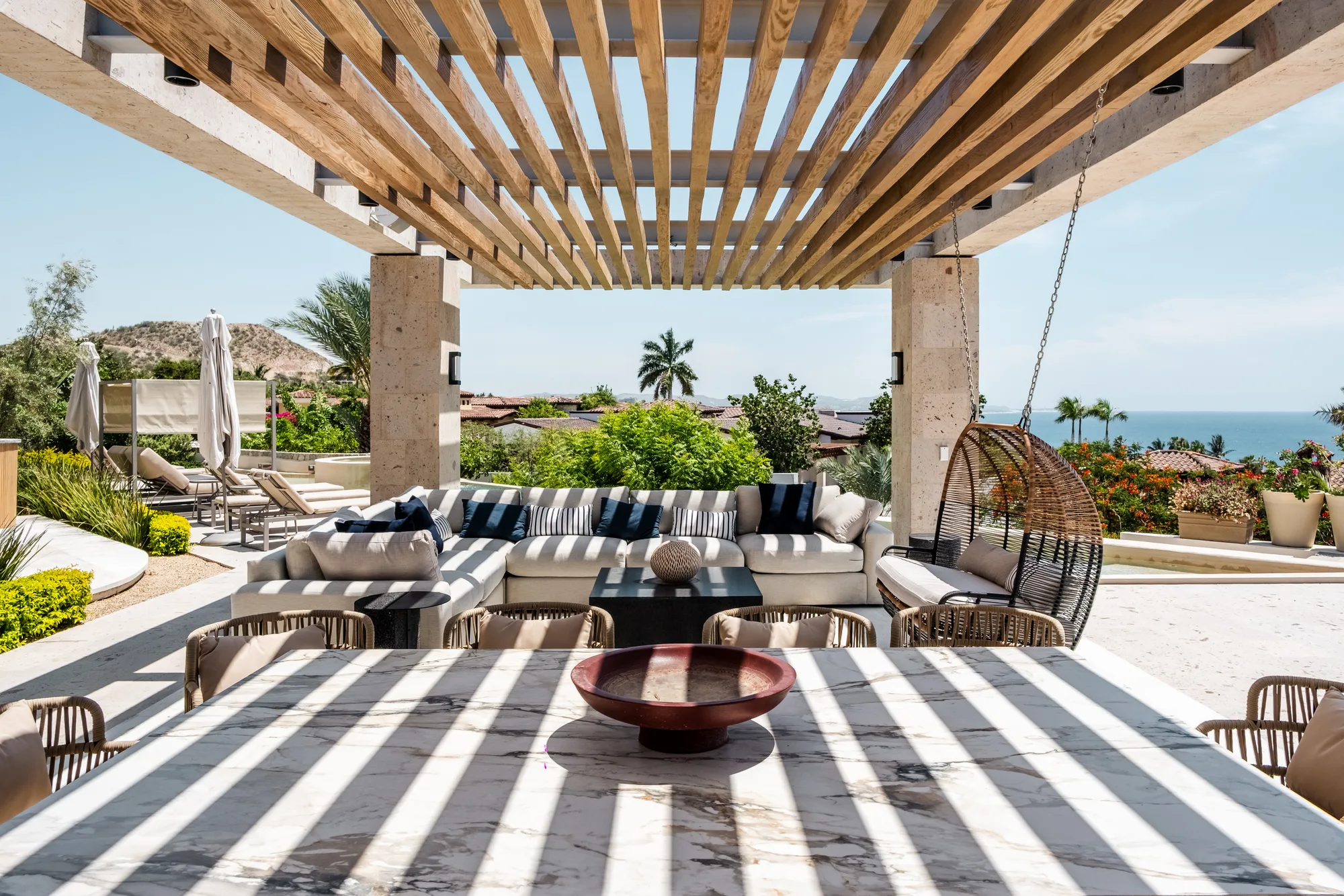 Shaded lounge terrace under a timber pergola with a sunken sofa and hanging chair, opening to palms and the sea