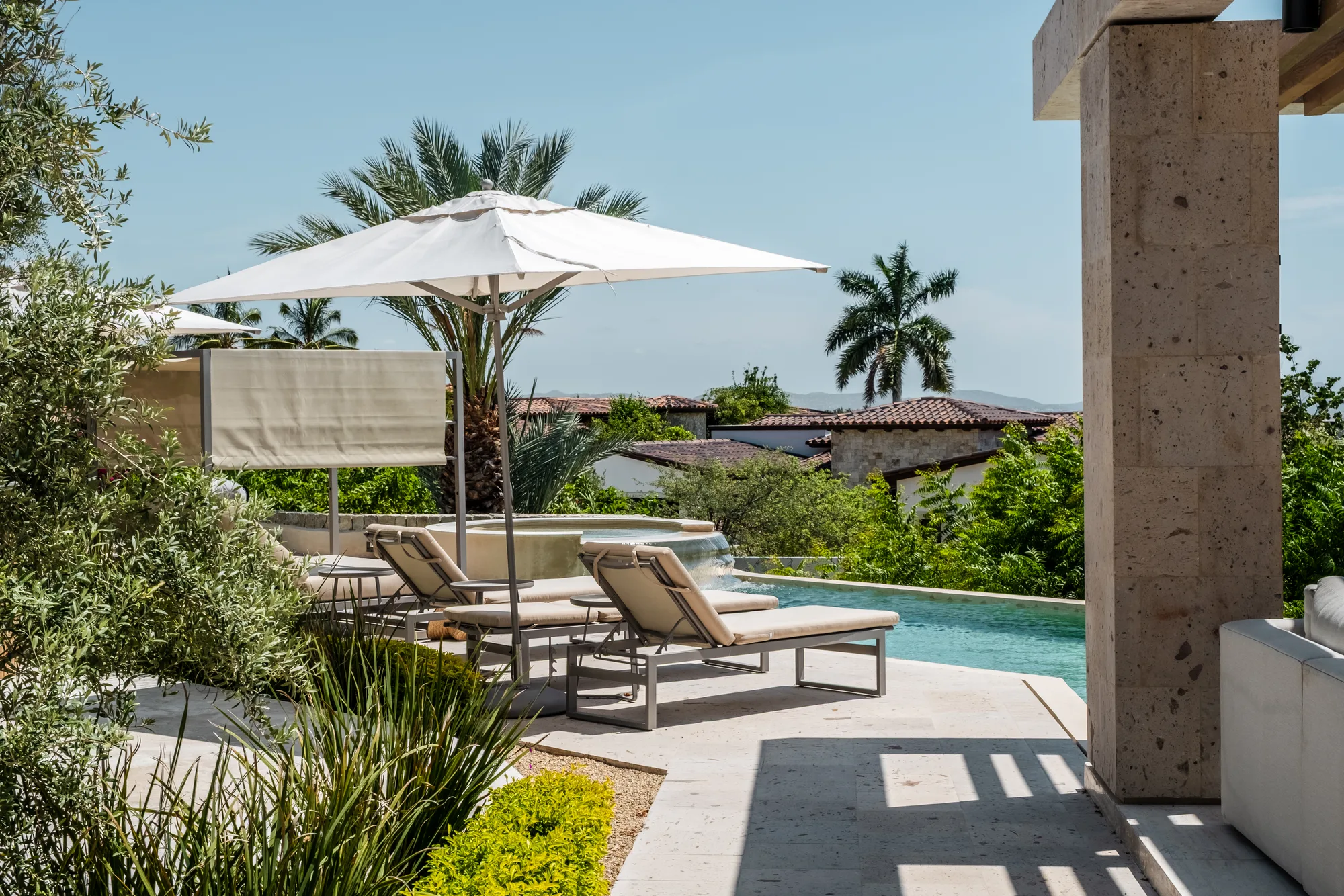 Pool terrace with loungers and a white umbrella beside a travertine column, a palm and hillside villas behind