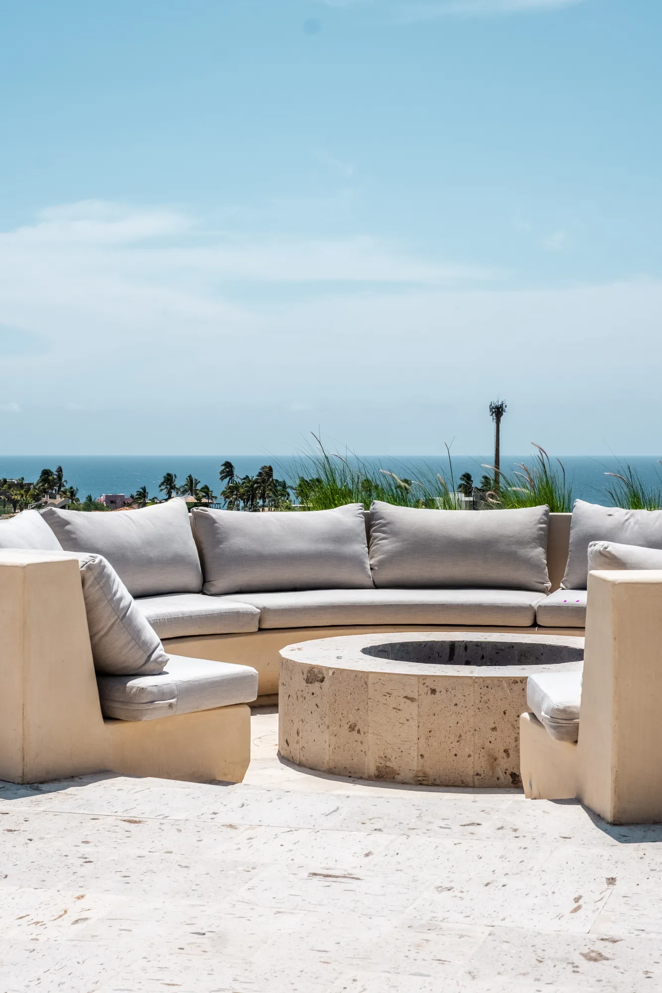 Rooftop lounge with a curved built-in sofa around a round travertine fire pit, facing the sea and palms