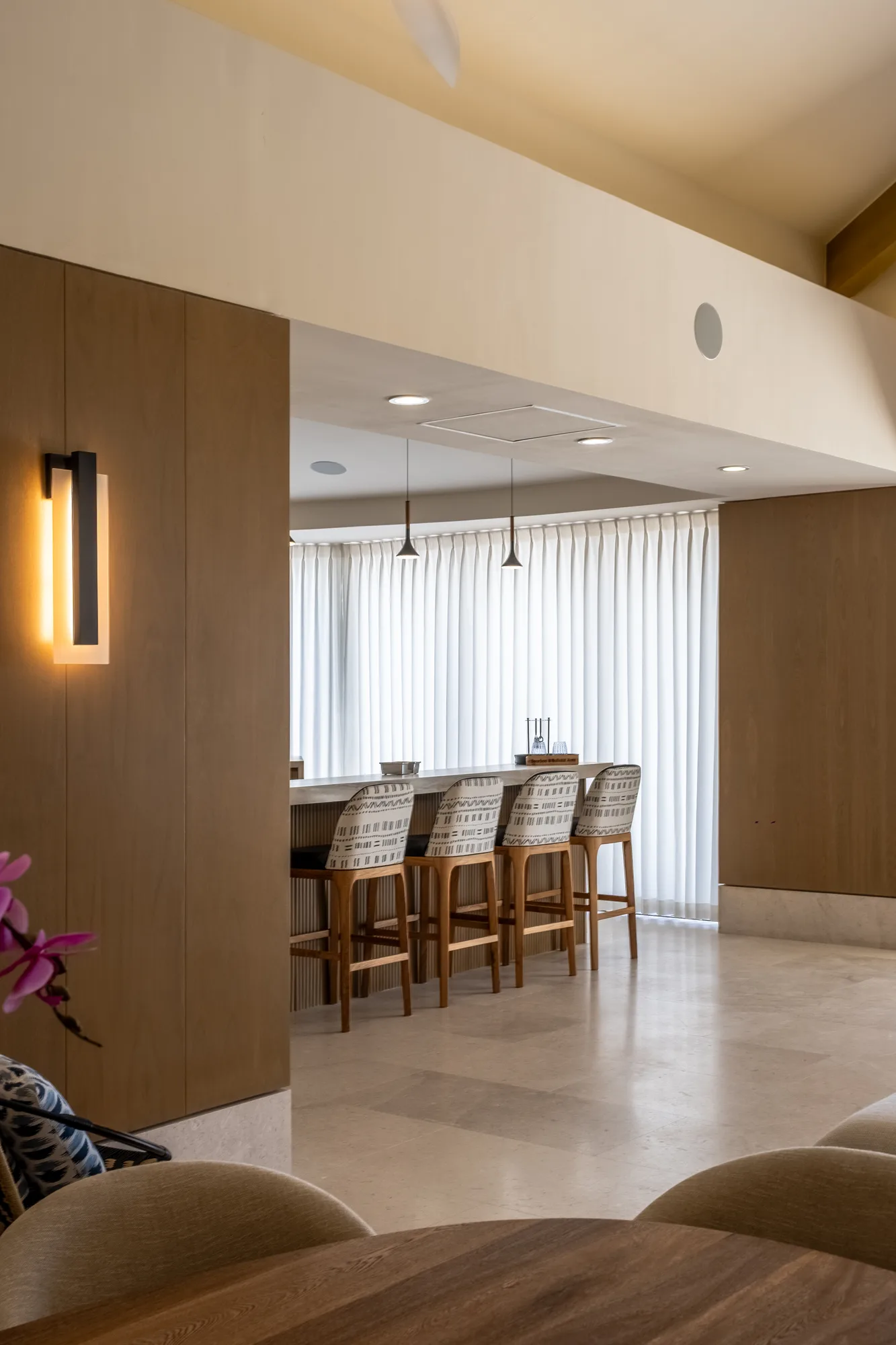 Interior bar with a marble counter and woven-leather stools against wood paneling, a lit sconce and sheer curtains