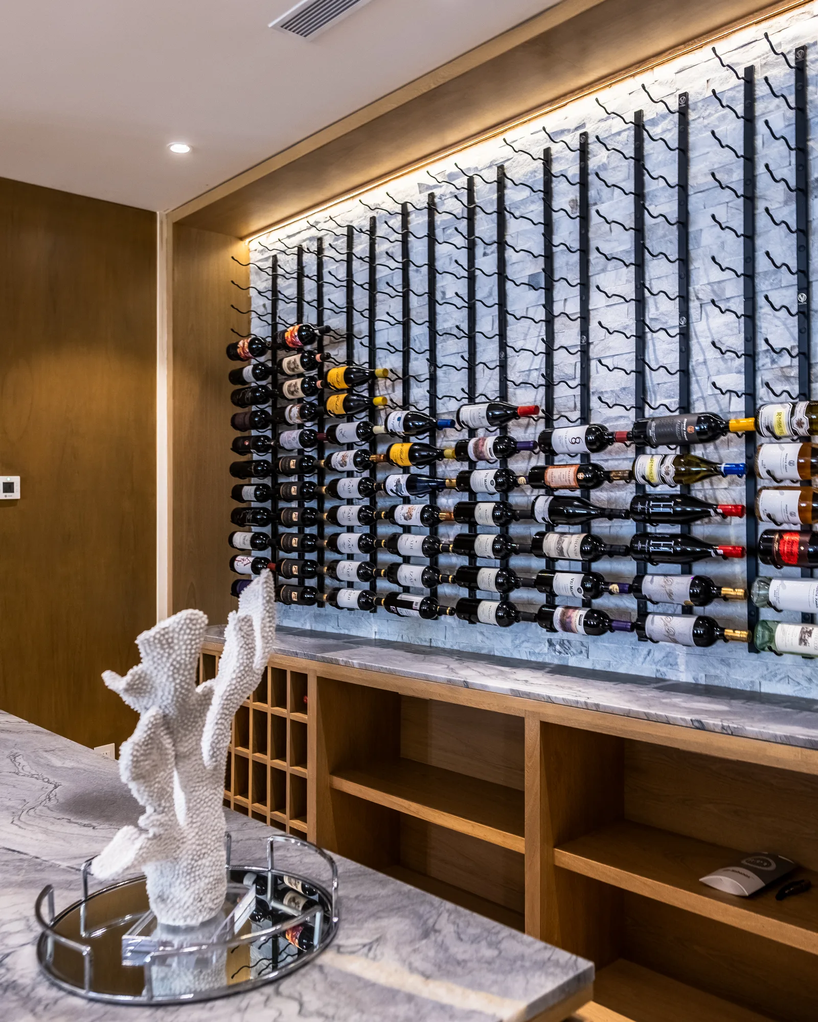 Backlit wine wall of black metal racks on marble, filled with bottles above wood cabinetry, with a coral sculpture