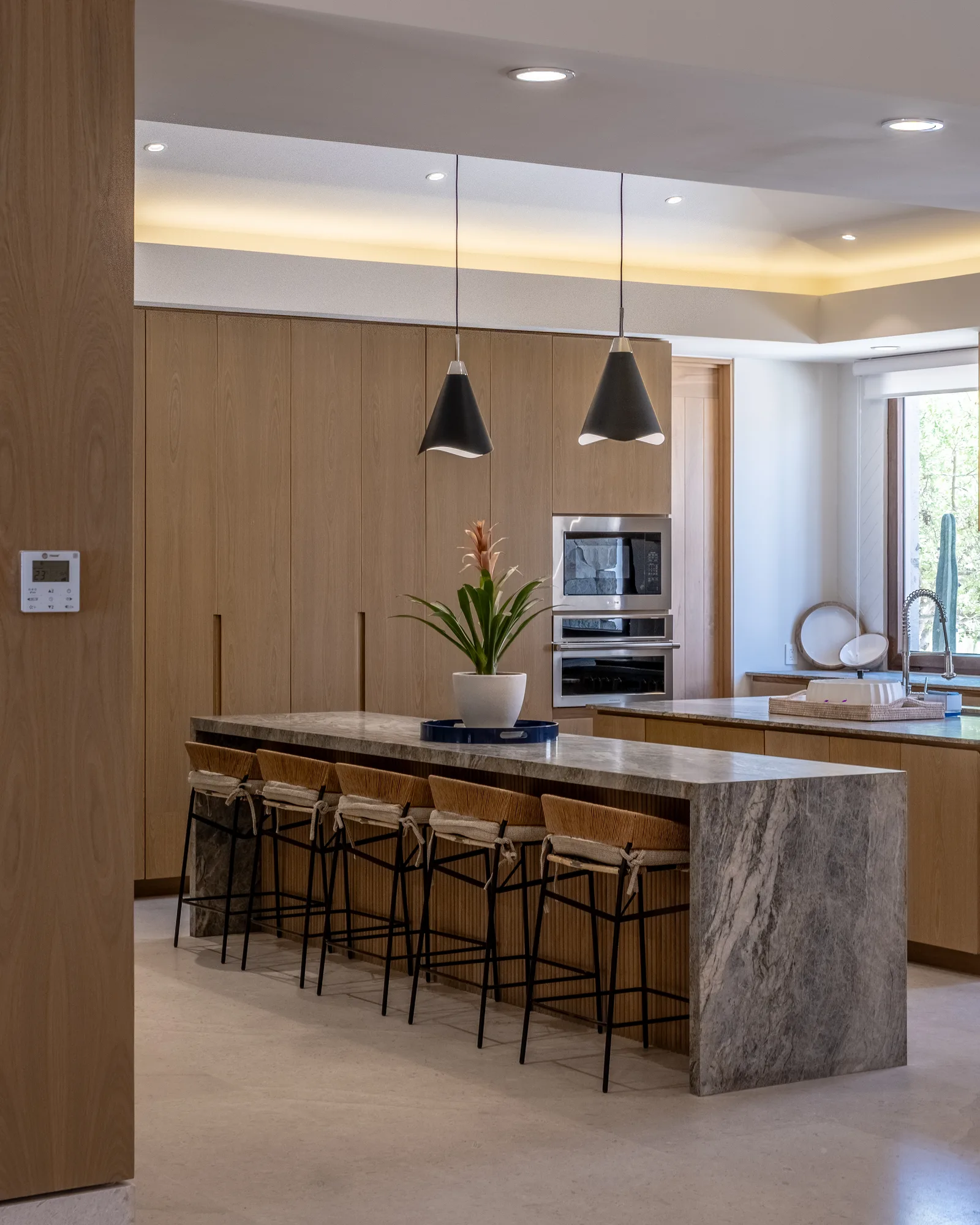 Kitchen with a grey marble waterfall island, woven stools and wood cabinetry under black pendant lights