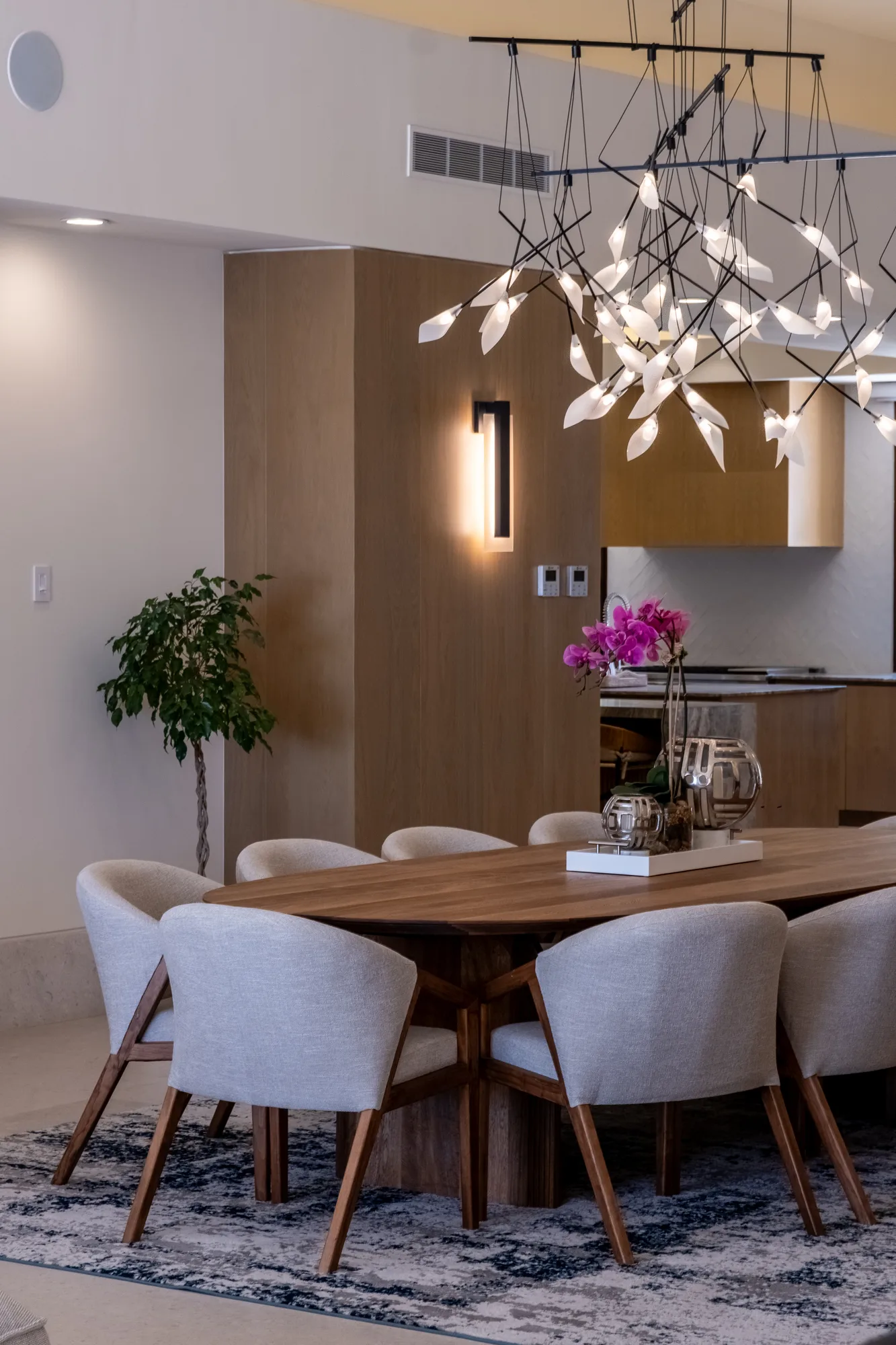 Dining room with an oval wood table and cream chairs under a branching light sculpture, dressed with orchids