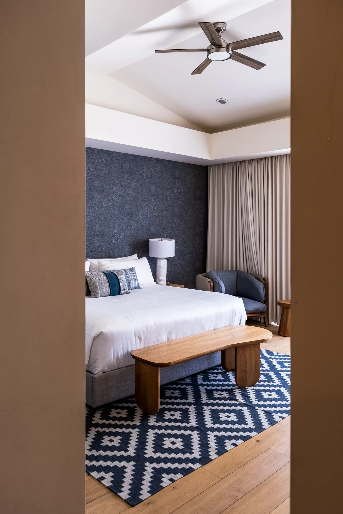 Primary bedroom with a patterned headboard wall, white linens, a curved wood bench and a diamond-motif rug