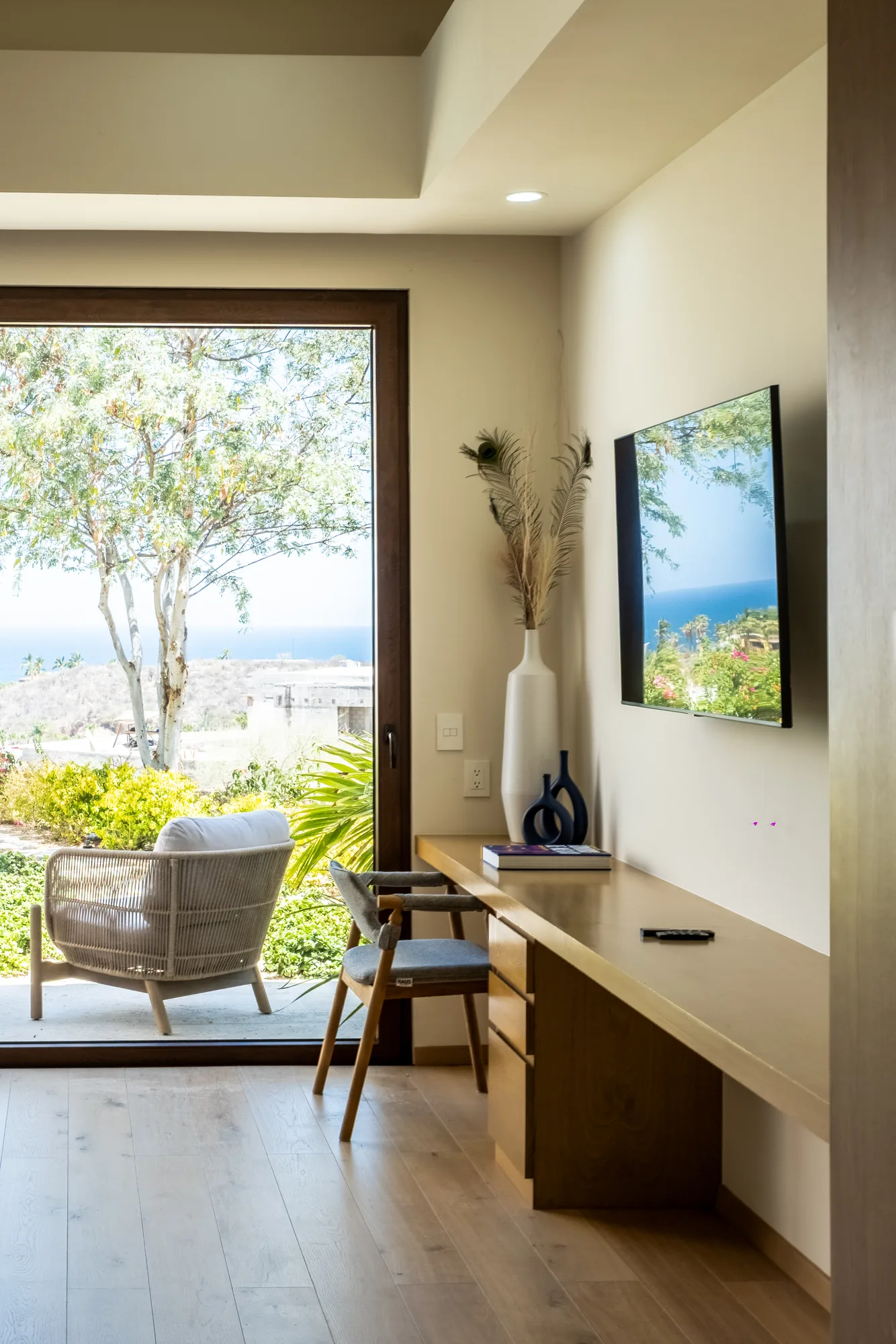 Bedroom desk nook by a floor-to-ceiling window with an ocean view, a woven armchair and pampas grass