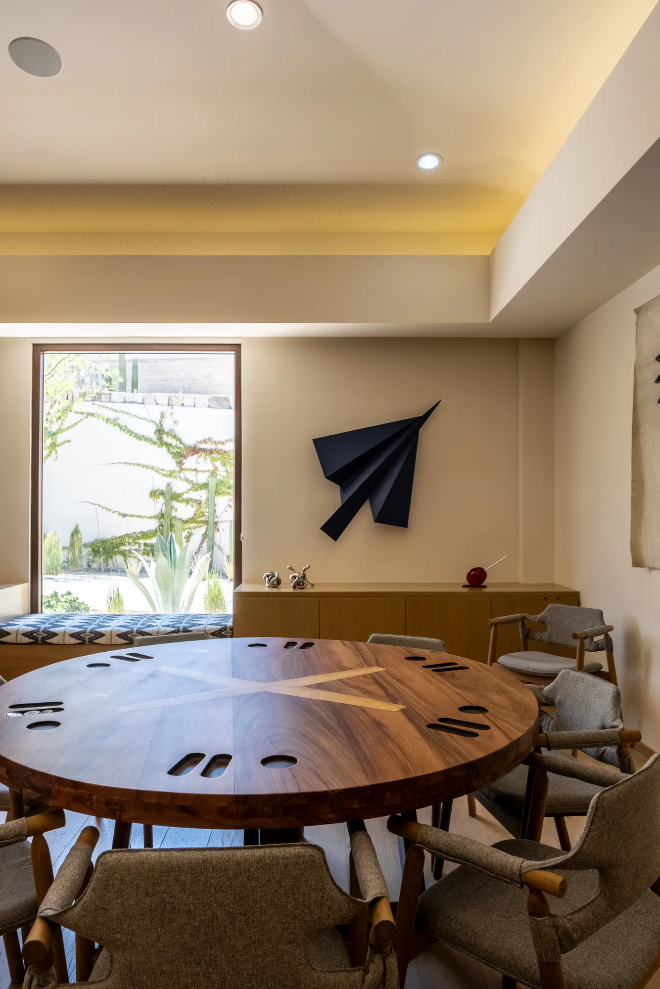 Breakfast area with a round walnut table and upholstered chairs beside a folded metal wall sculpture and a cactus-garden window