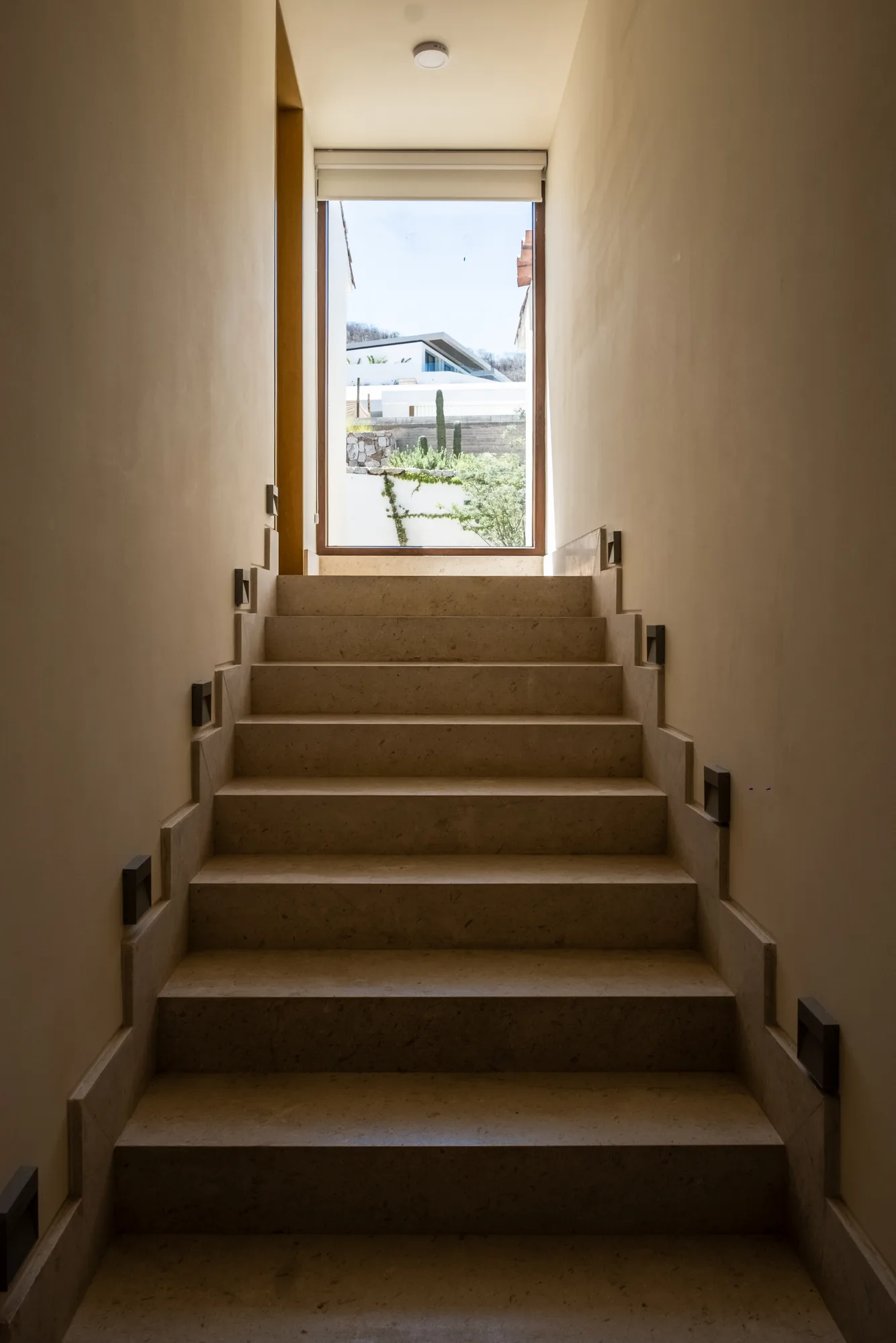 Travertine staircase rising toward a window that frames a neighboring villa and cacti, with recessed step lights