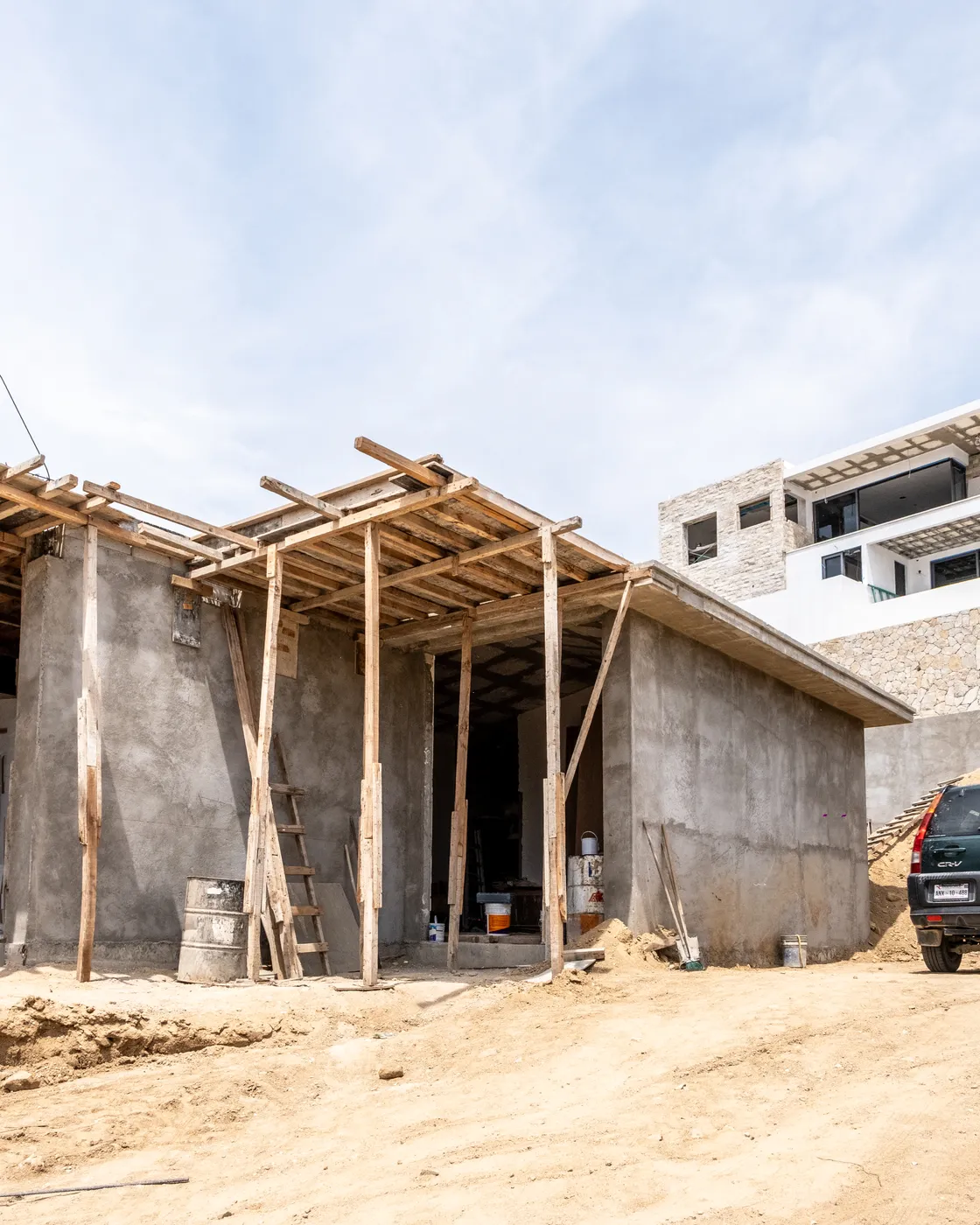 Concrete shell of the house with timber props supporting a cantilevered slab, a finished villa rising beyond on the site