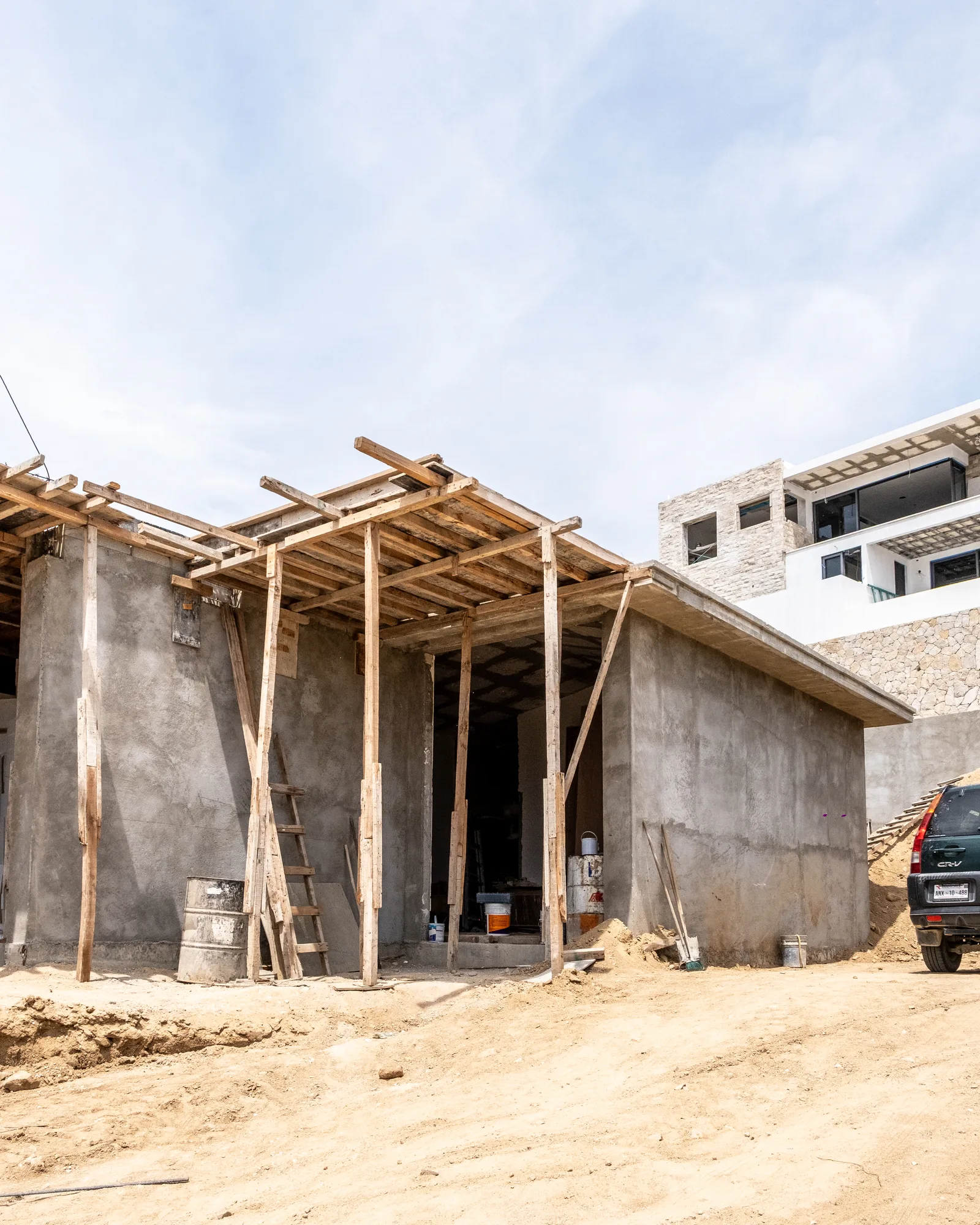 Concrete shell of the house with timber props supporting a cantilevered slab, a finished villa rising beyond on the site