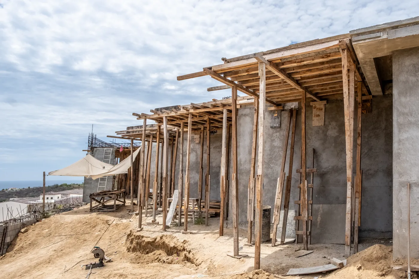 Row of concrete rooms under timber-shored slab overhangs on a hillside site, with a shade sail and the sea beyond