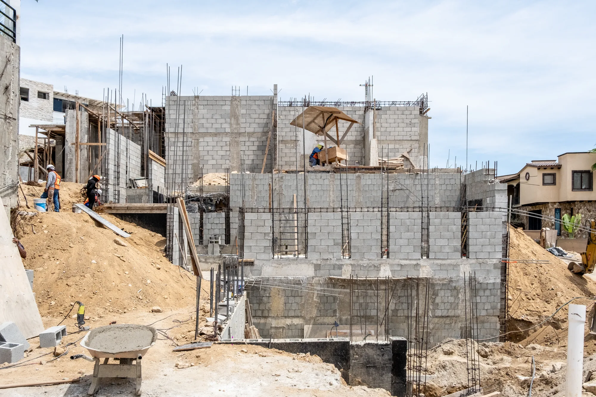 Wider construction site with rising block walls, rebar columns and workers, neighboring houses behind