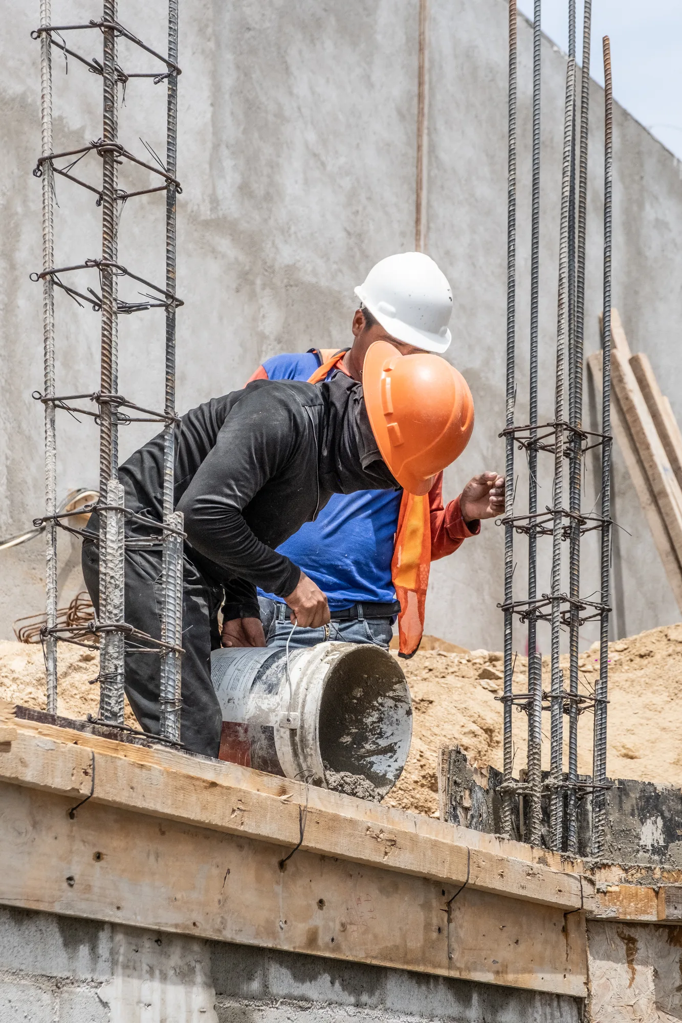 Two workers in hard hats placing concrete around rebar columns against a poured wall
