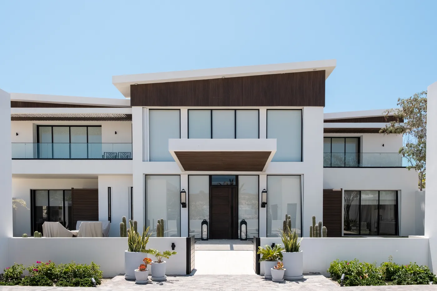 Symmetrical white modern facade with dark wood cladding, full-height glass and a cantilevered entry canopy, Chileno RE 30 in Los Cabos