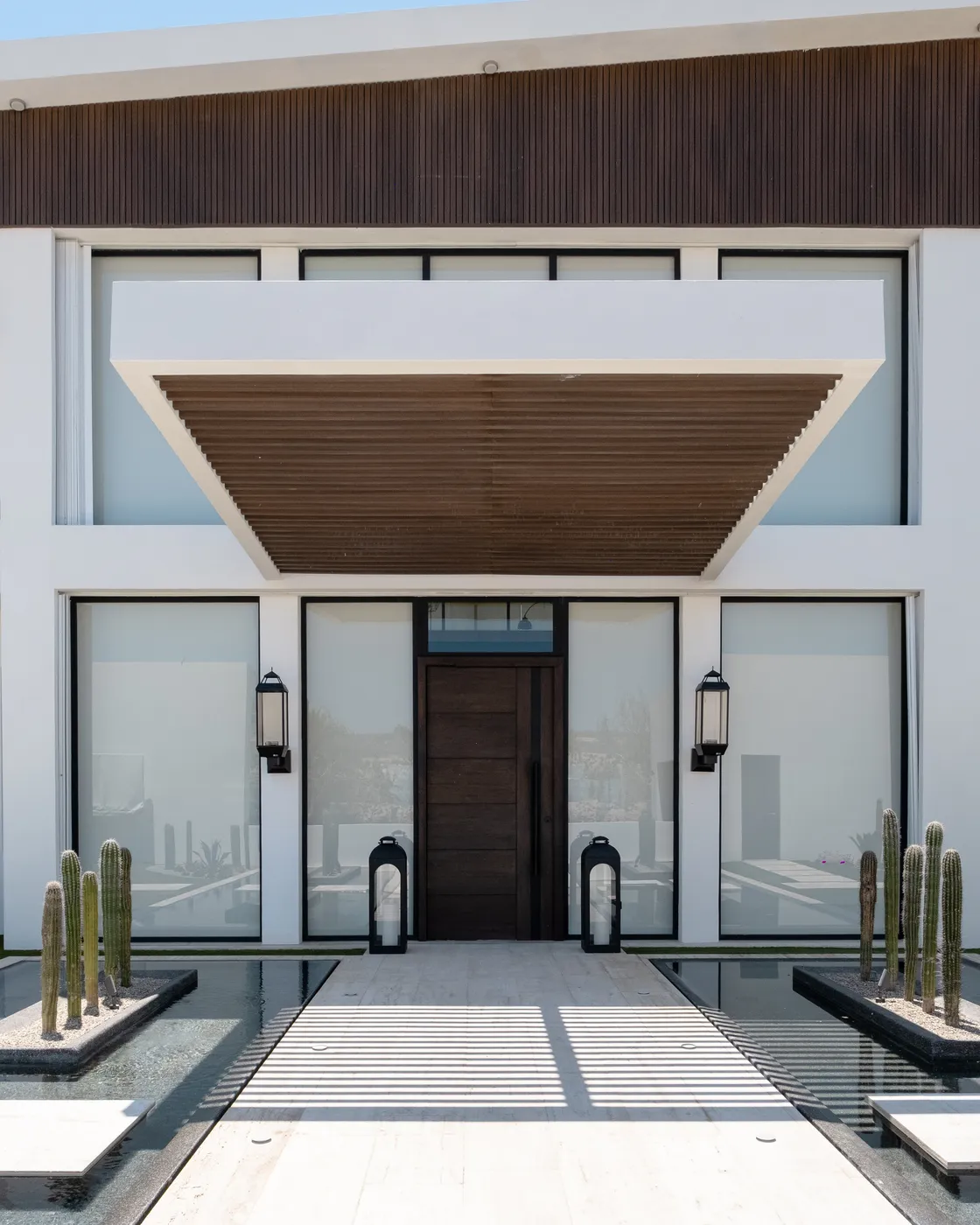 Dark wood front door under a slatted wood canopy between glass walls, flanked by reflecting pools and cacti