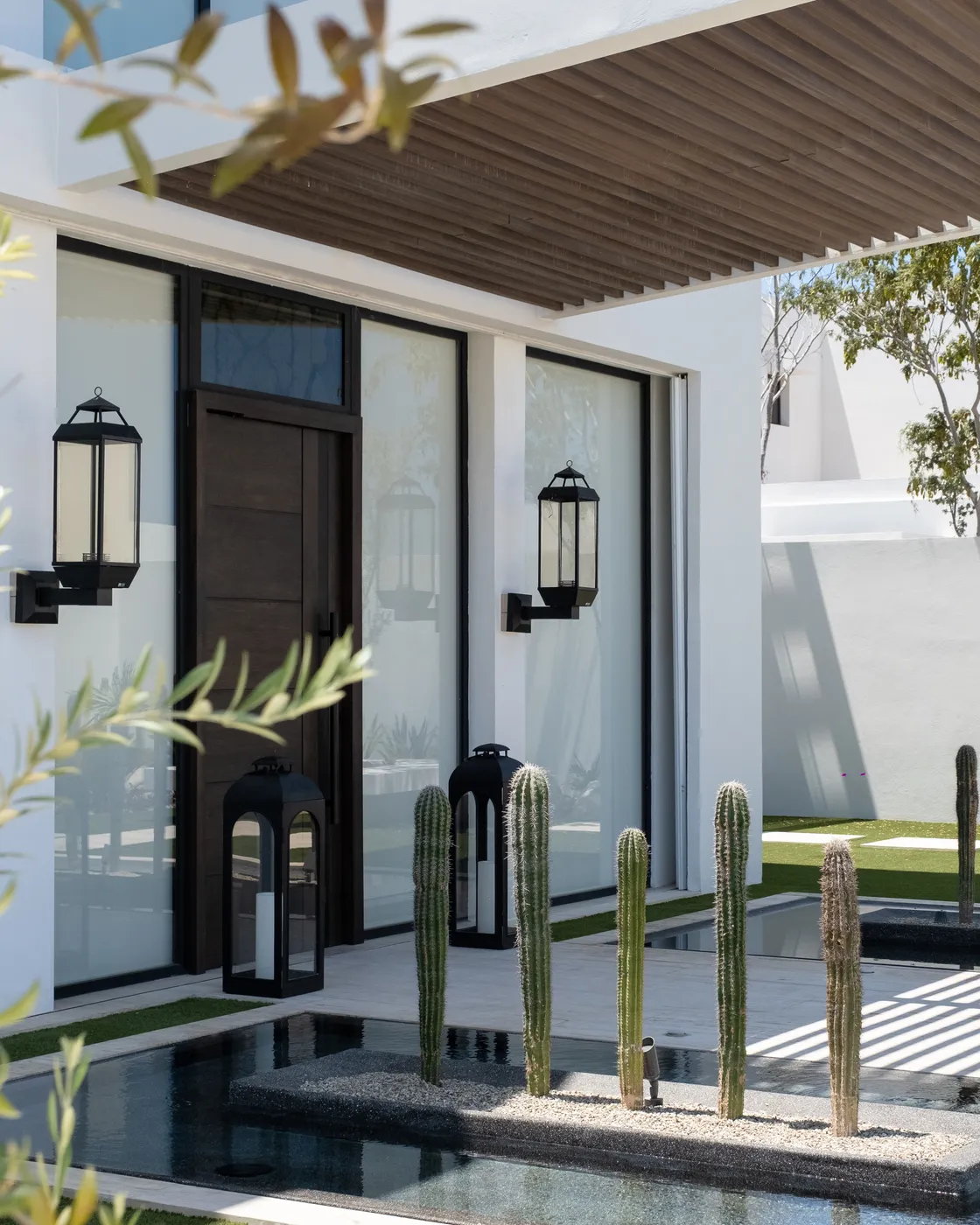 Entrance detail with a wood door and black lanterns under a wood-slat pergola beside a cactus-lined reflecting pool