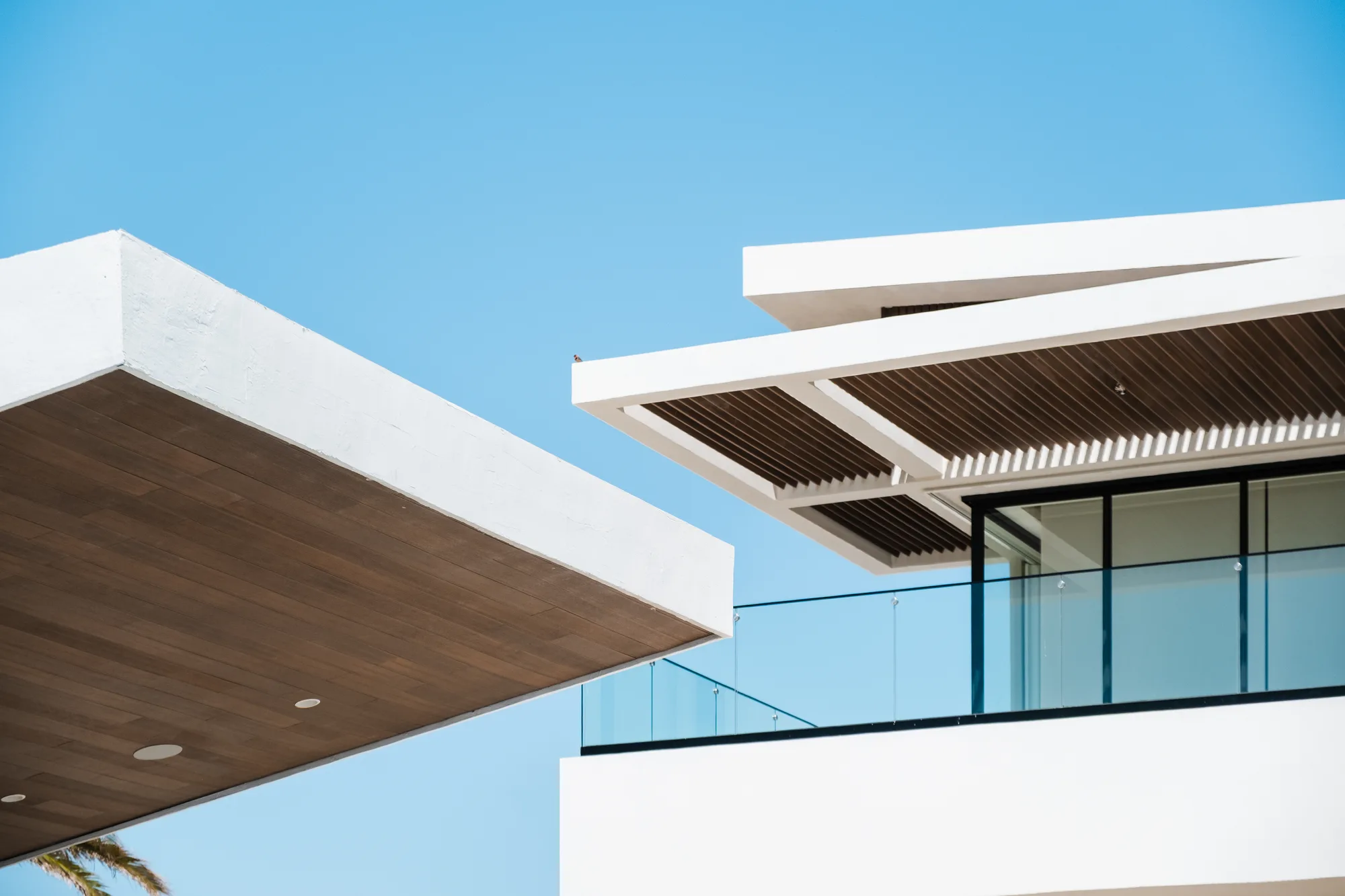 Detail of two cantilevered rooflines, a wood soffit and a slatted brise-soleil over a glass-railed balcony against the sky