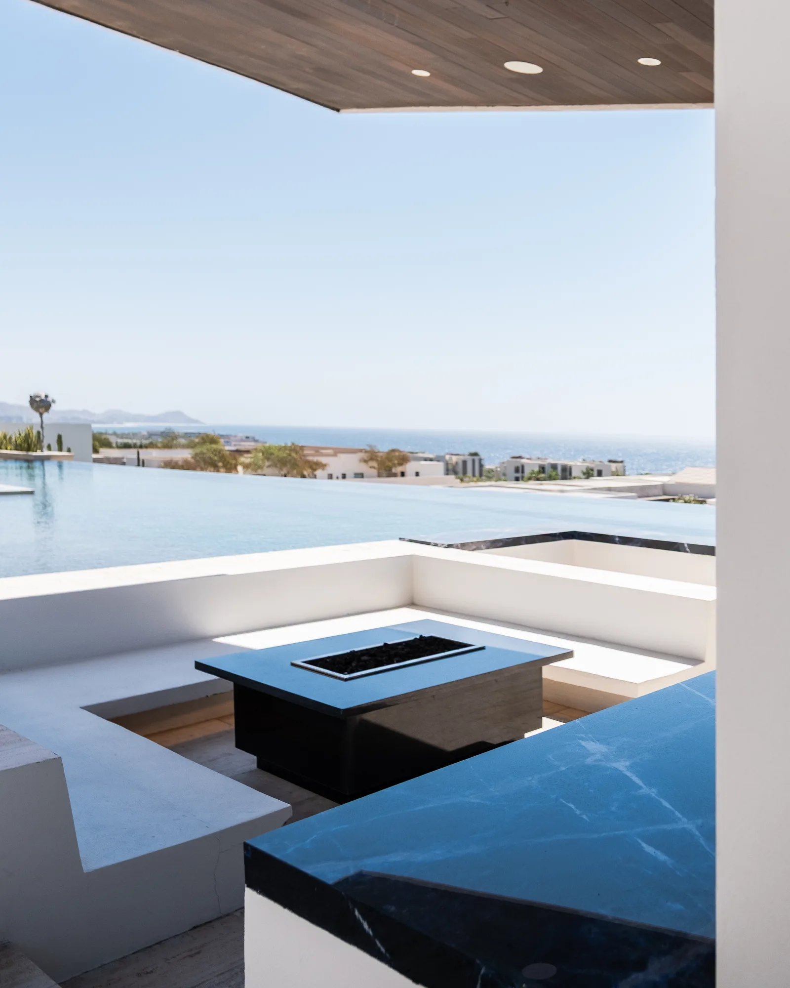 Sunken lounge with a black fire table under a covered terrace, opening past the infinity pool to the sea