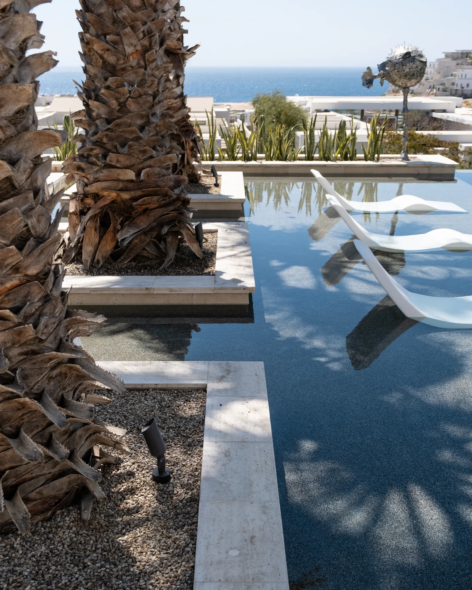 Poolside detail of date palm trunks and travertine stepping strips over a dark reflecting pool, the sea in the distance