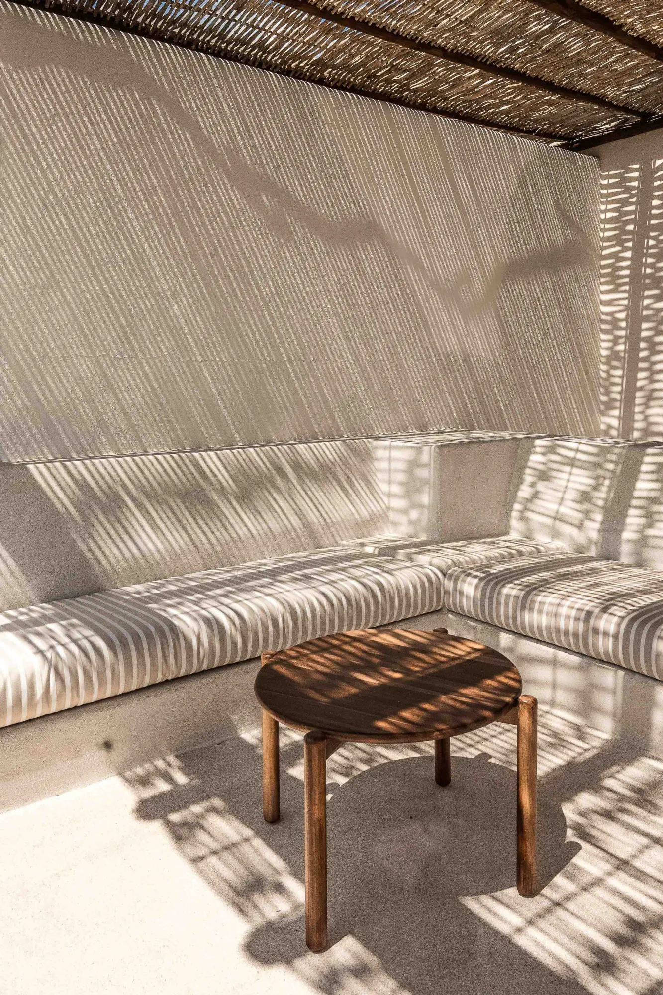 Shaded terrace where a reed roof casts striped light over plaster banquettes and a round wood table