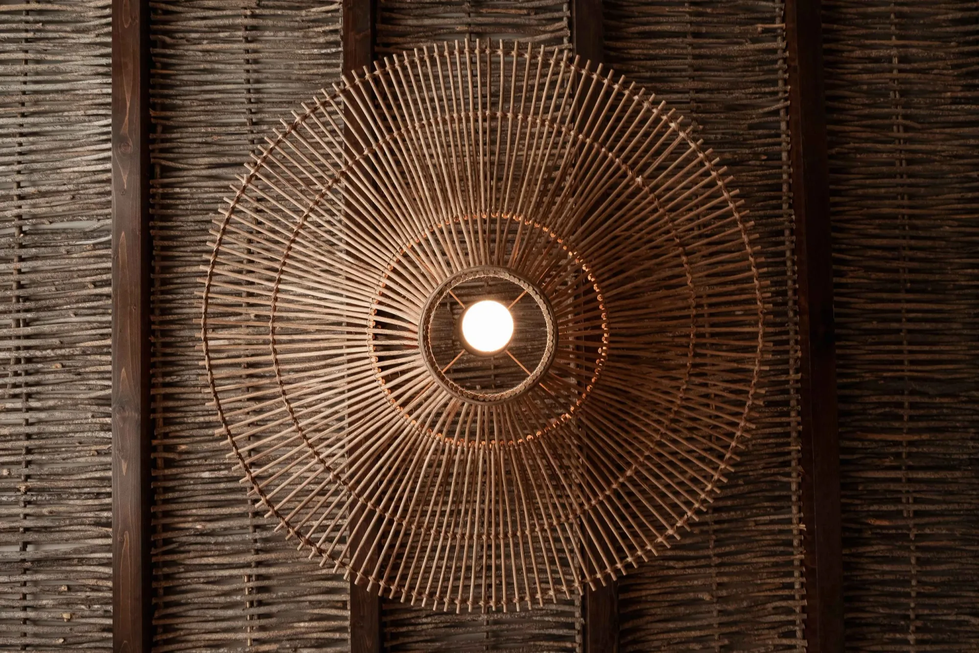 A round woven rattan pendant glowing against a dark reed-and-timber ceiling
