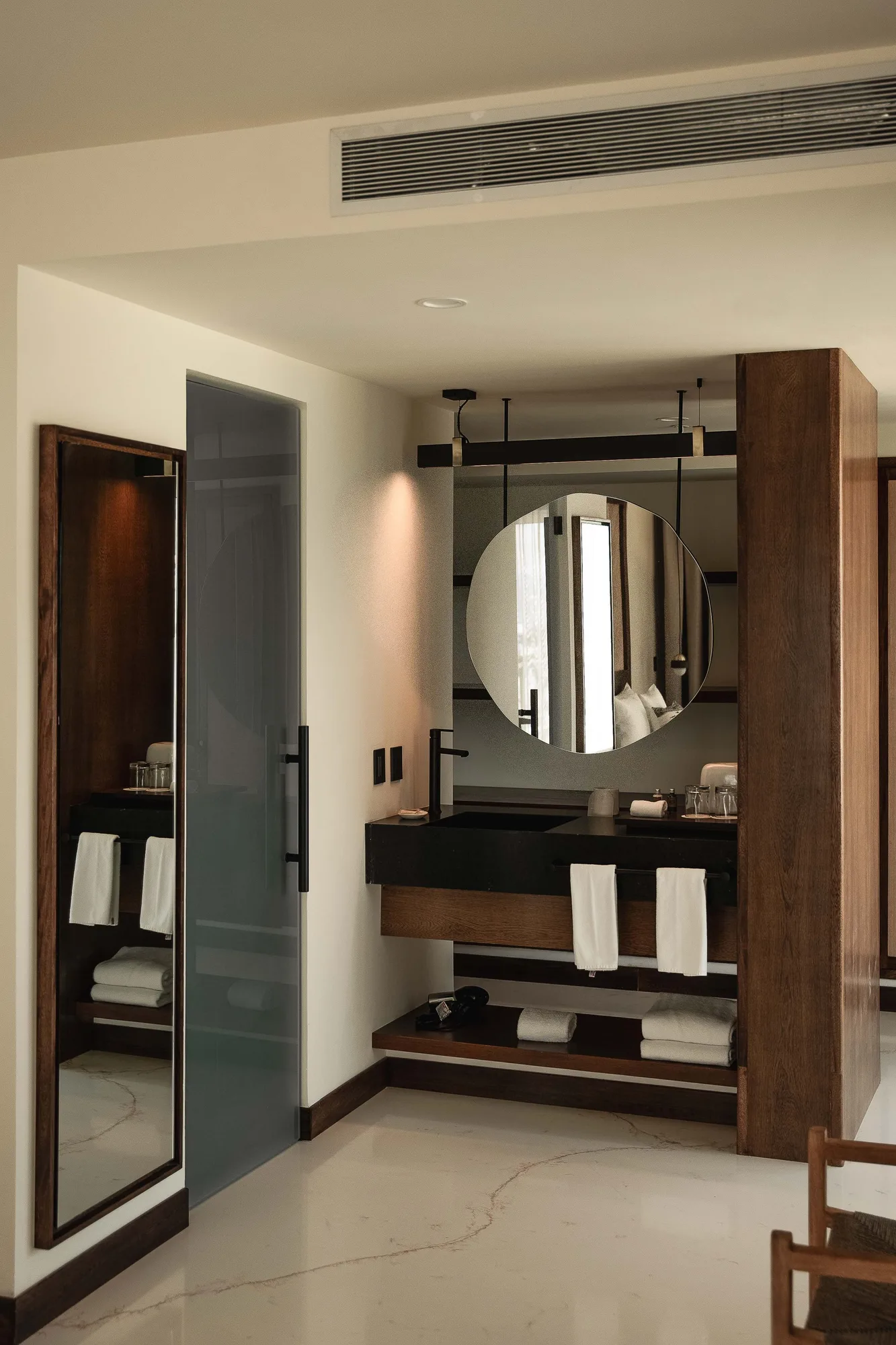 Dressing area with a round mirror, wood cabinetry, a frosted glass shower door and folded towels on marble