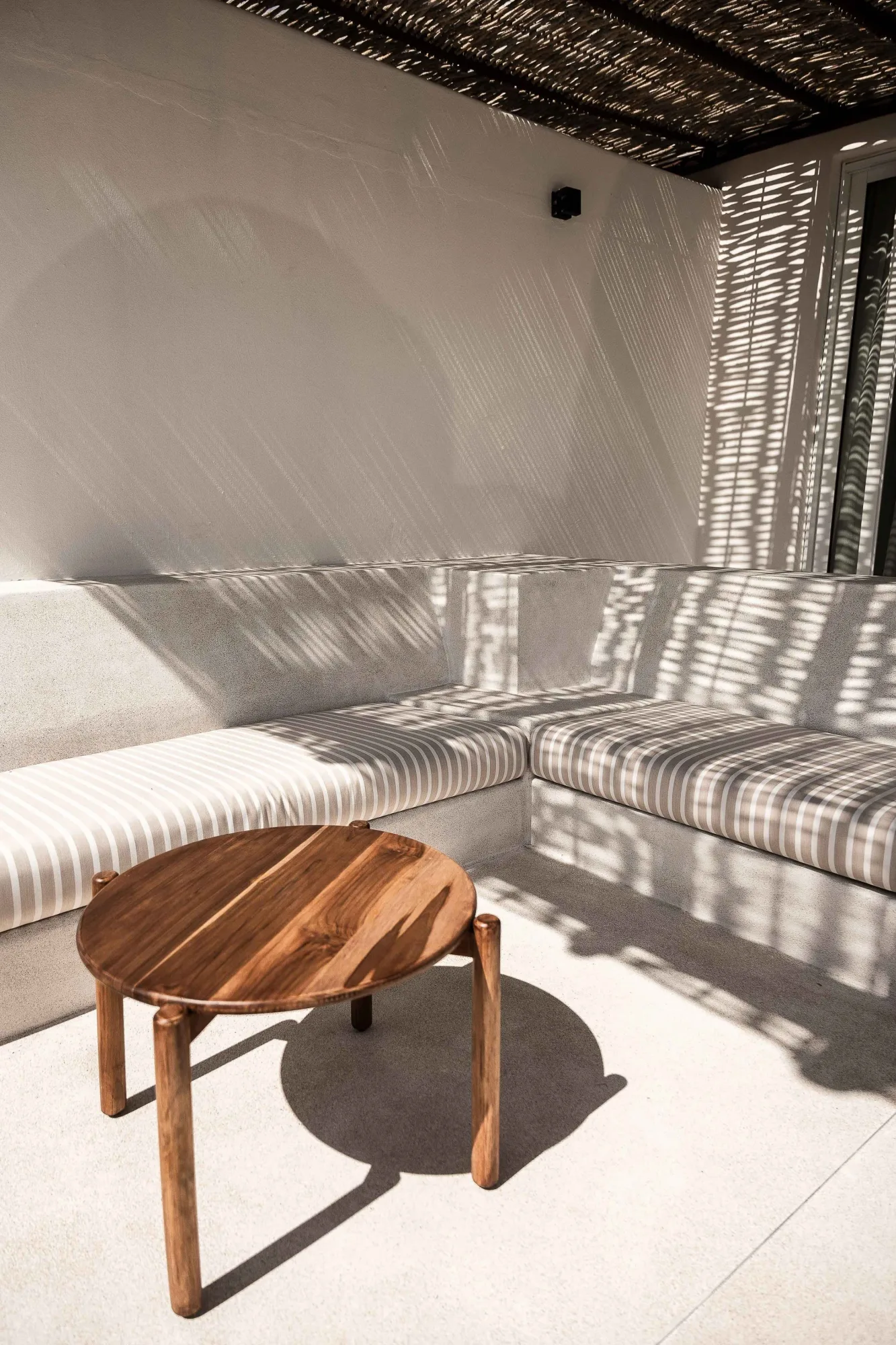 Shaded terrace corner with an L-shaped plaster banquette, striped cushions and a round wood table under a reed roof
