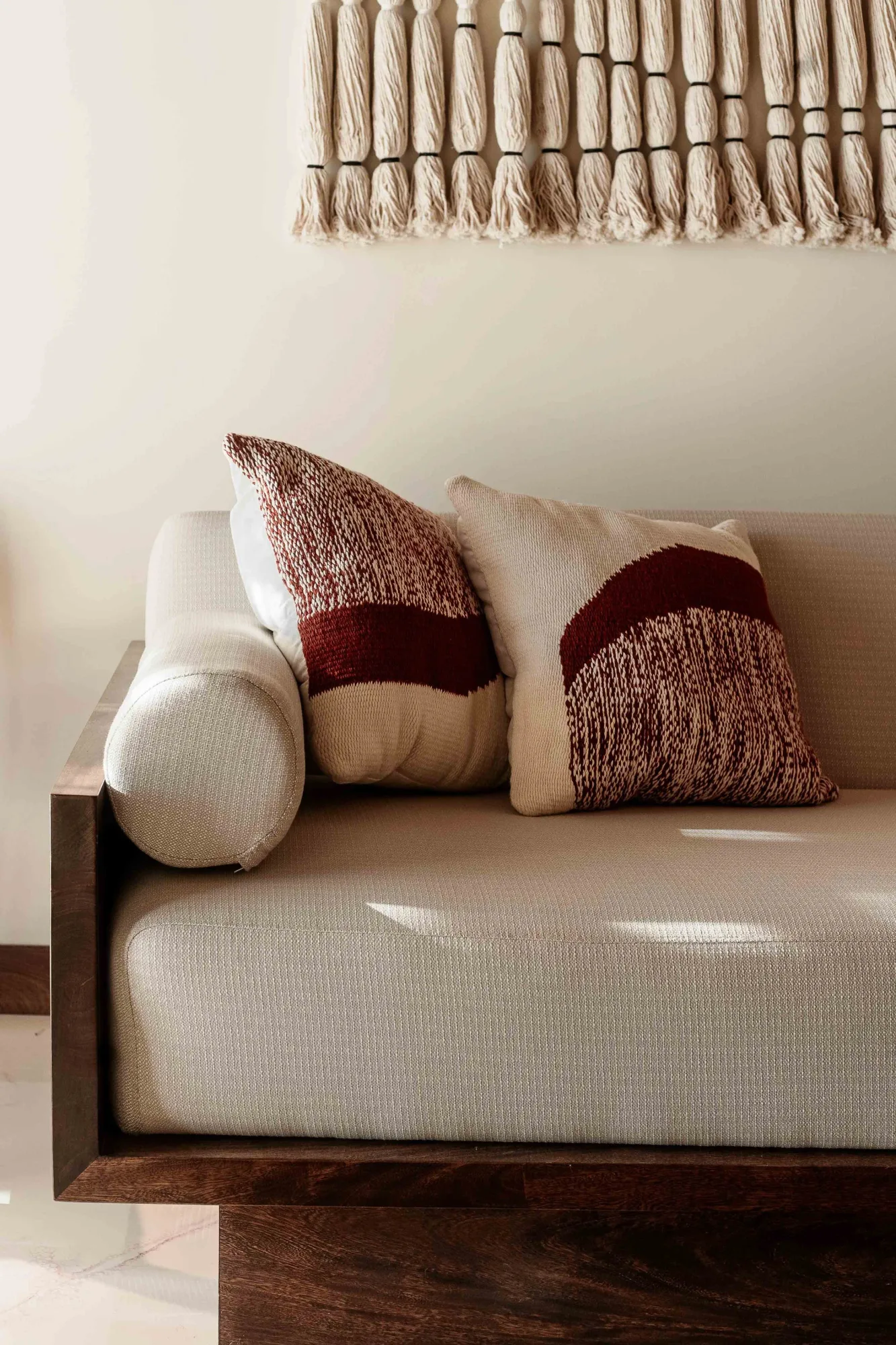 White sofa with a bolster and rust-and-cream cushions under a fringed macramé wall hanging