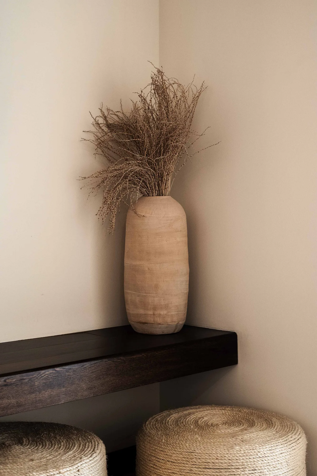 Vignette of a pale turned-wood vase with dried grasses on a dark floating shelf above woven jute stools