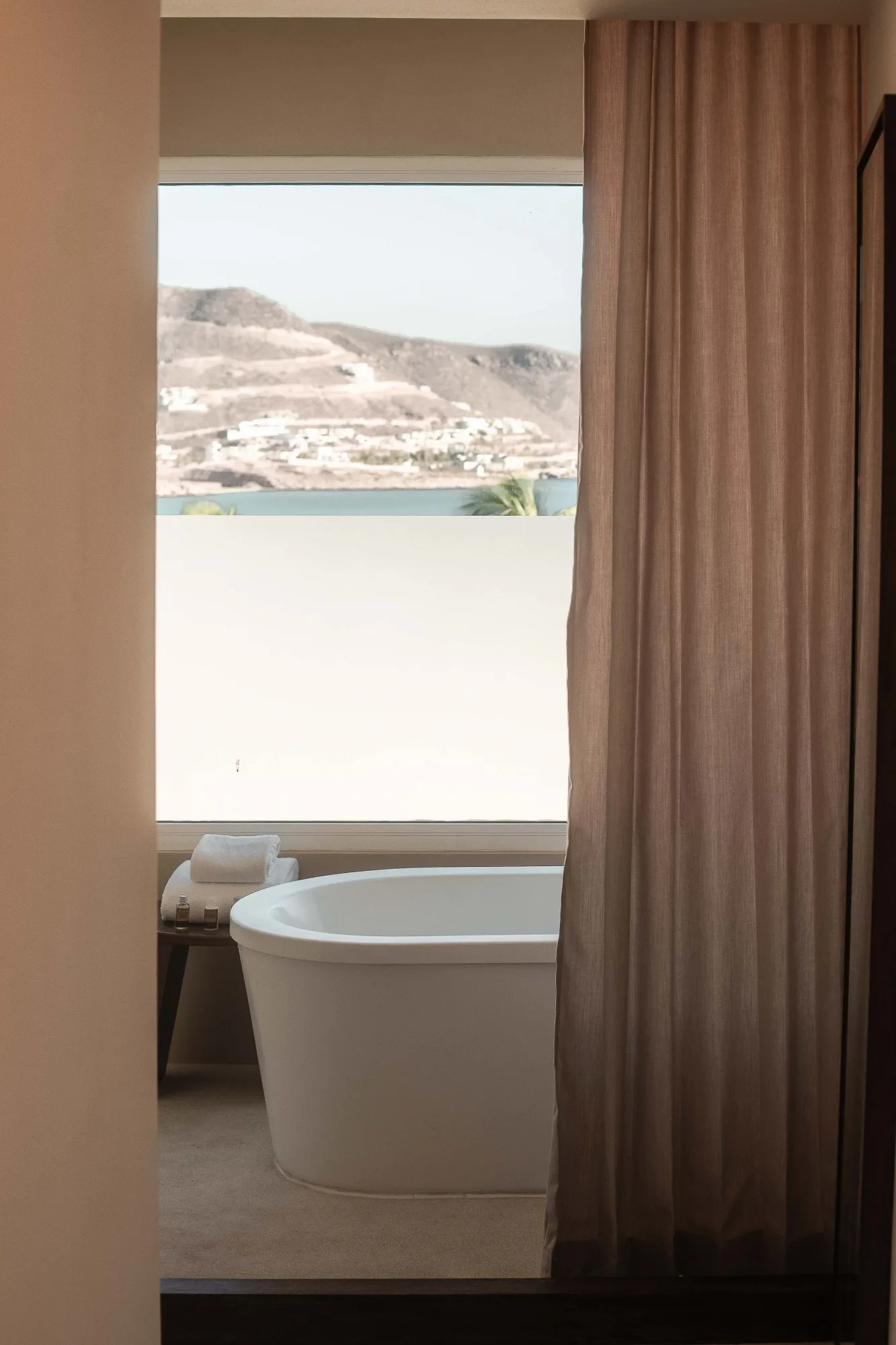 Freestanding white soaking tub at a window framing the bay, mountains and marina, towels on a wood bench, Hotel Indigo in La Paz