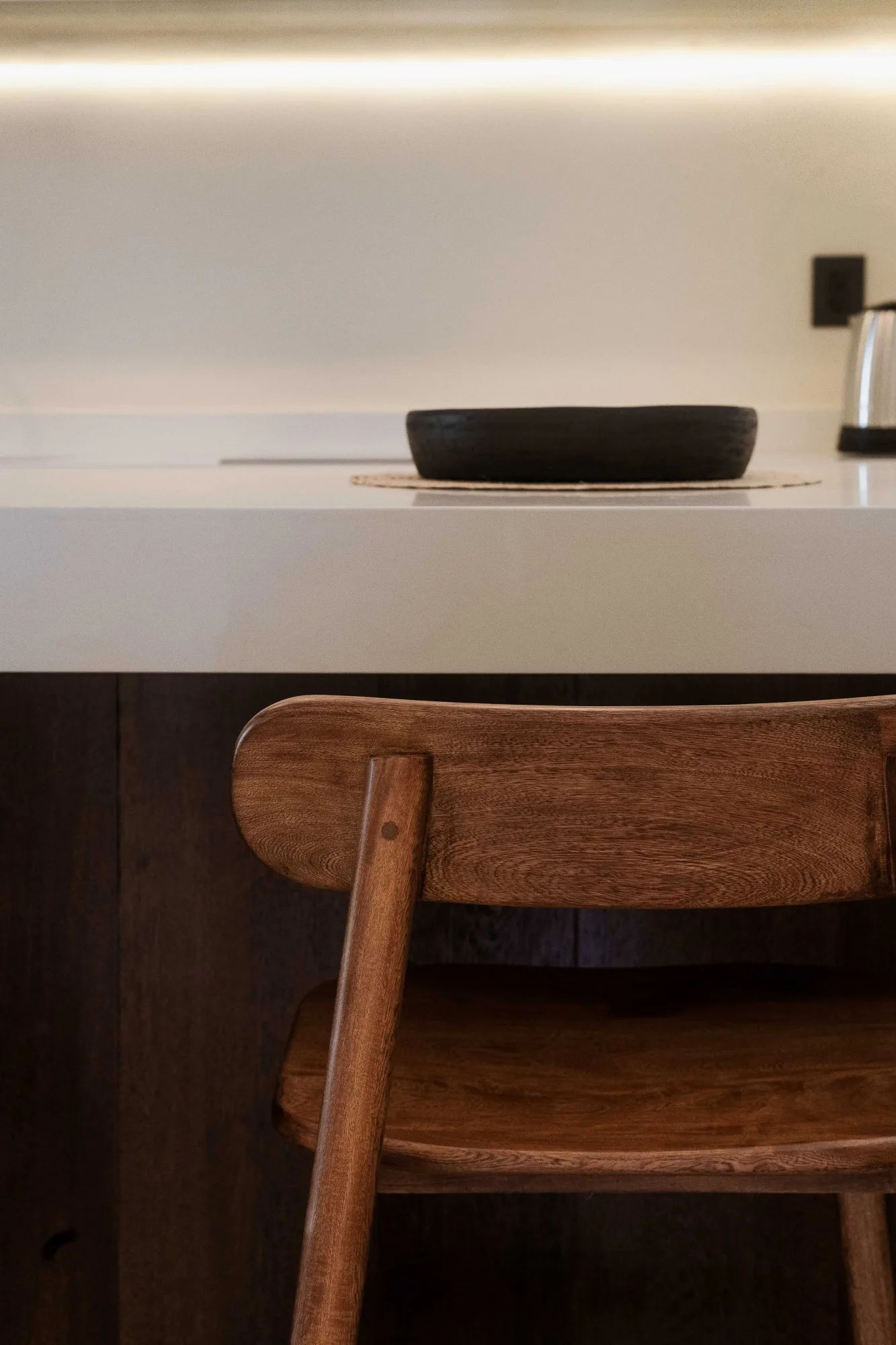 Table styling with a black bowl on a woven mat on a pale stone counter, a wood chair back in the foreground