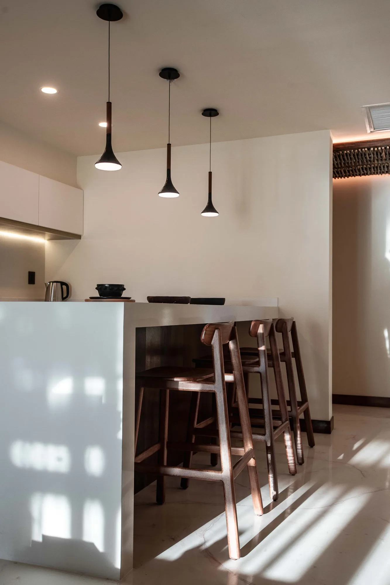 Suite bar counter with three cone pendants and walnut stools, sunlight patterning the polished floor