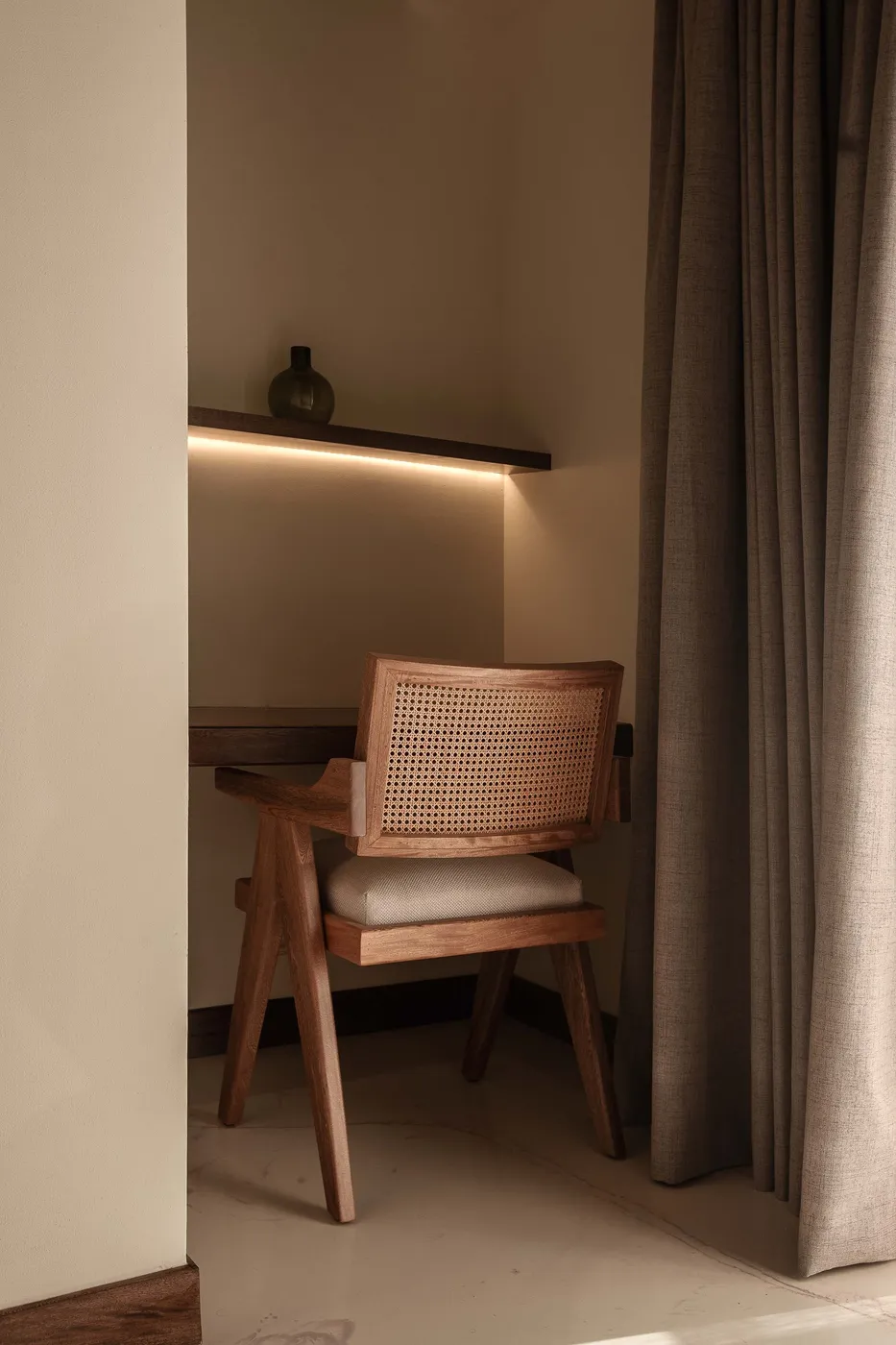 Desk nook with a cane-back wood chair at a floating desk beneath a lit shelf holding a small vessel