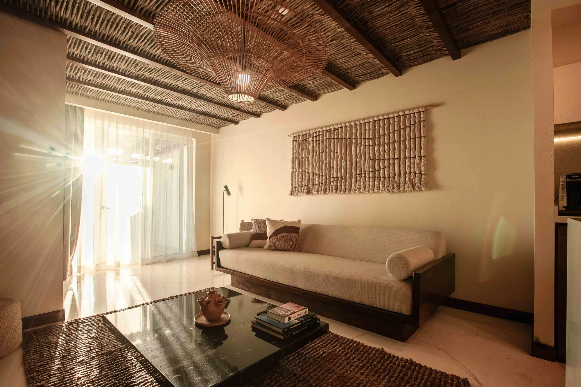 Bright suite lounge with a reed-and-timber ceiling, a large rattan pendant, white sofa and macramé hanging, light flaring through sheers