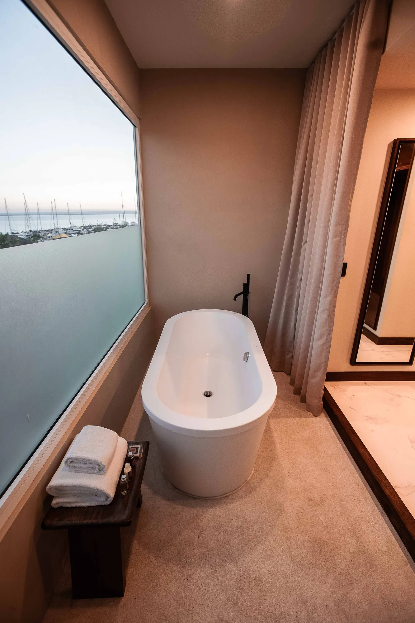 Freestanding tub at a large window with a marina and sea view, towels on a wood bench and a tall mirror at dusk