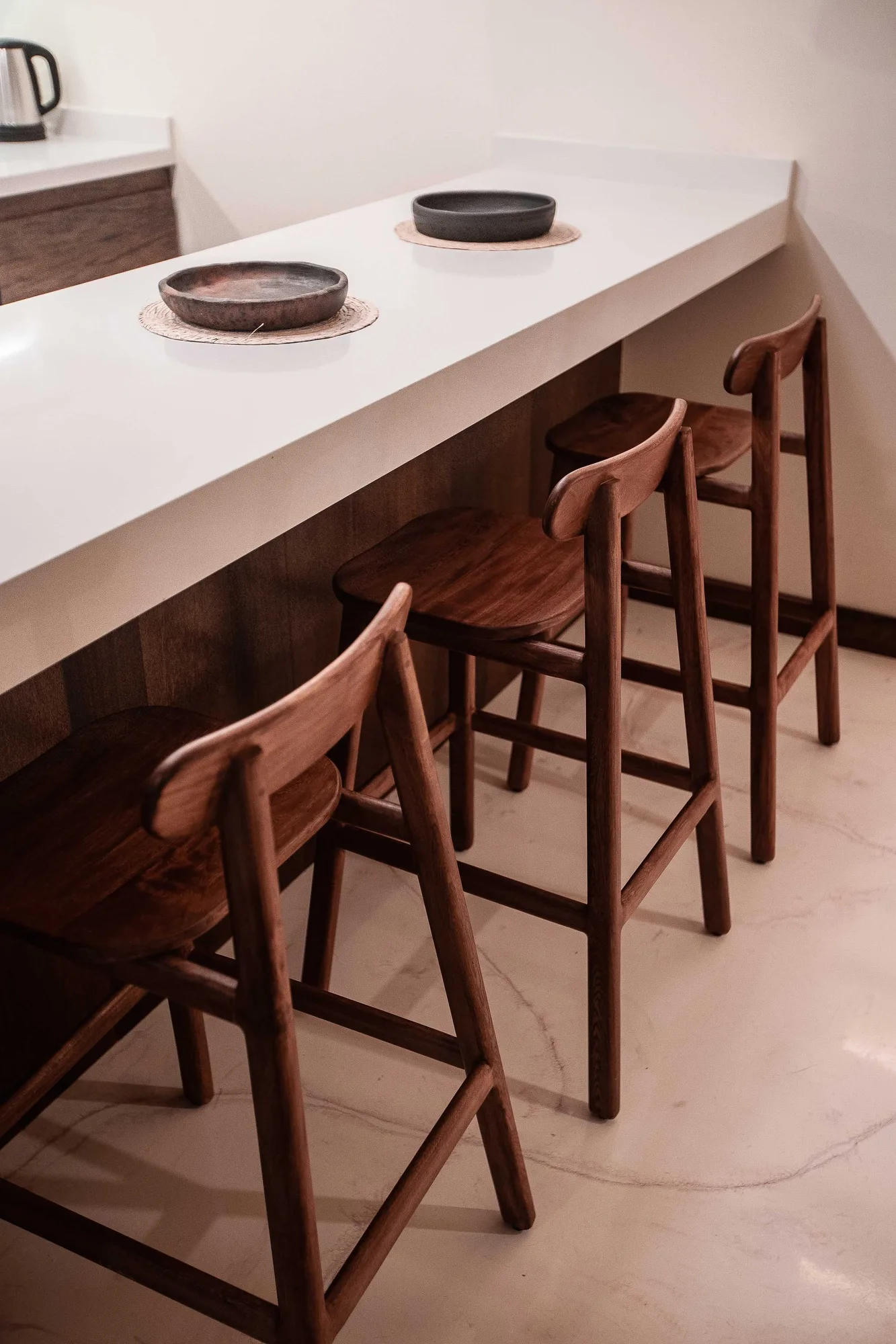 Three walnut stools tucked under a white waterfall counter with shallow bowls on woven mats