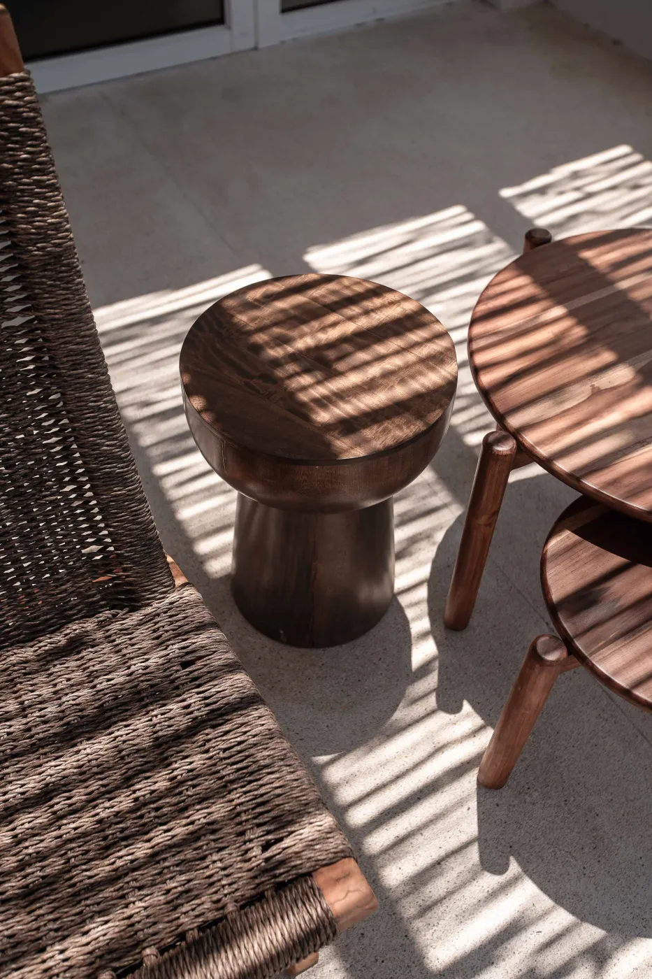 An hourglass walnut stool and a round wood table on a terrace under striped light, a woven chair alongside