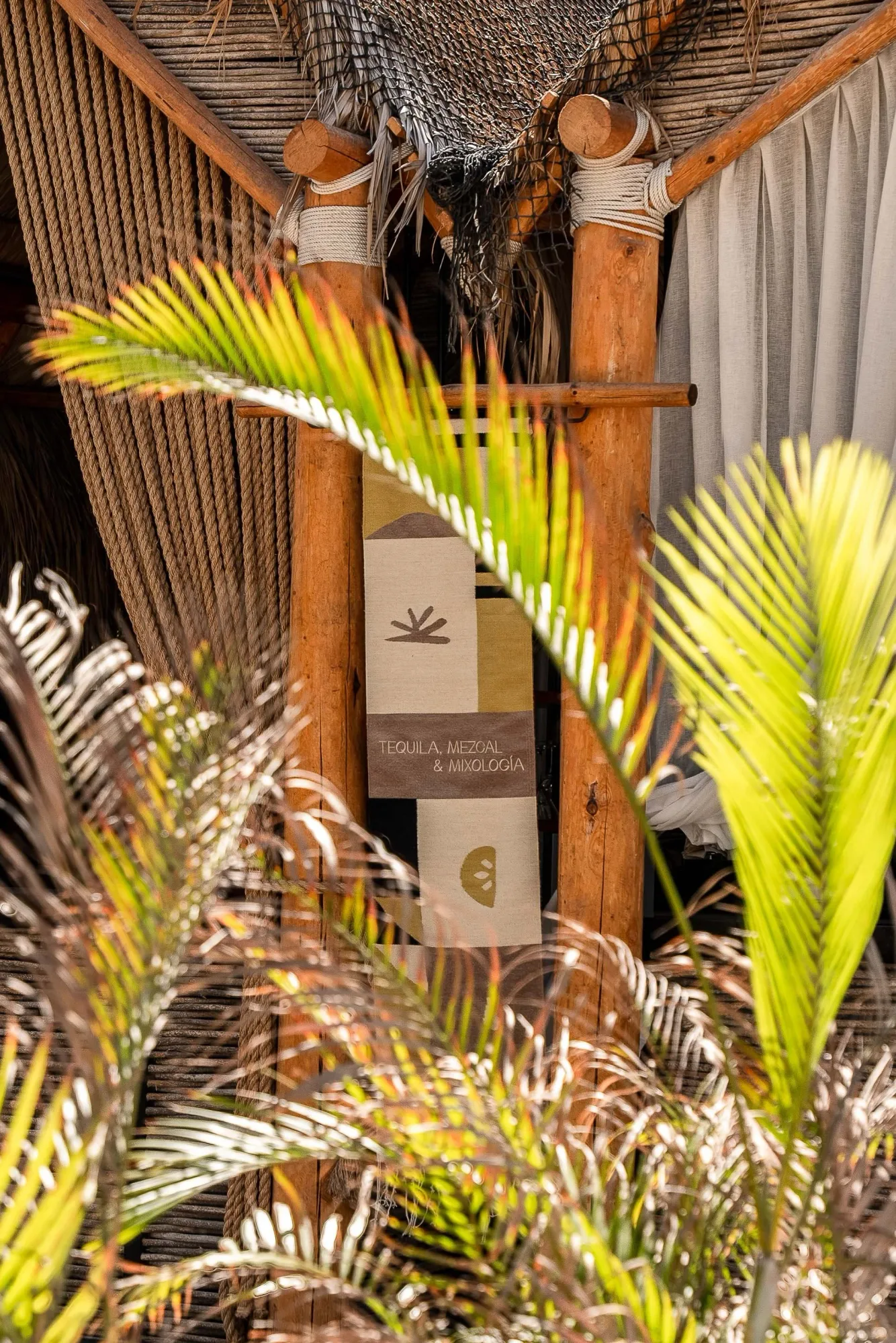 Branding detail of a printed mixology menu hanging on a rope-lashed timber post of the palapa bar, framed by palm fronds