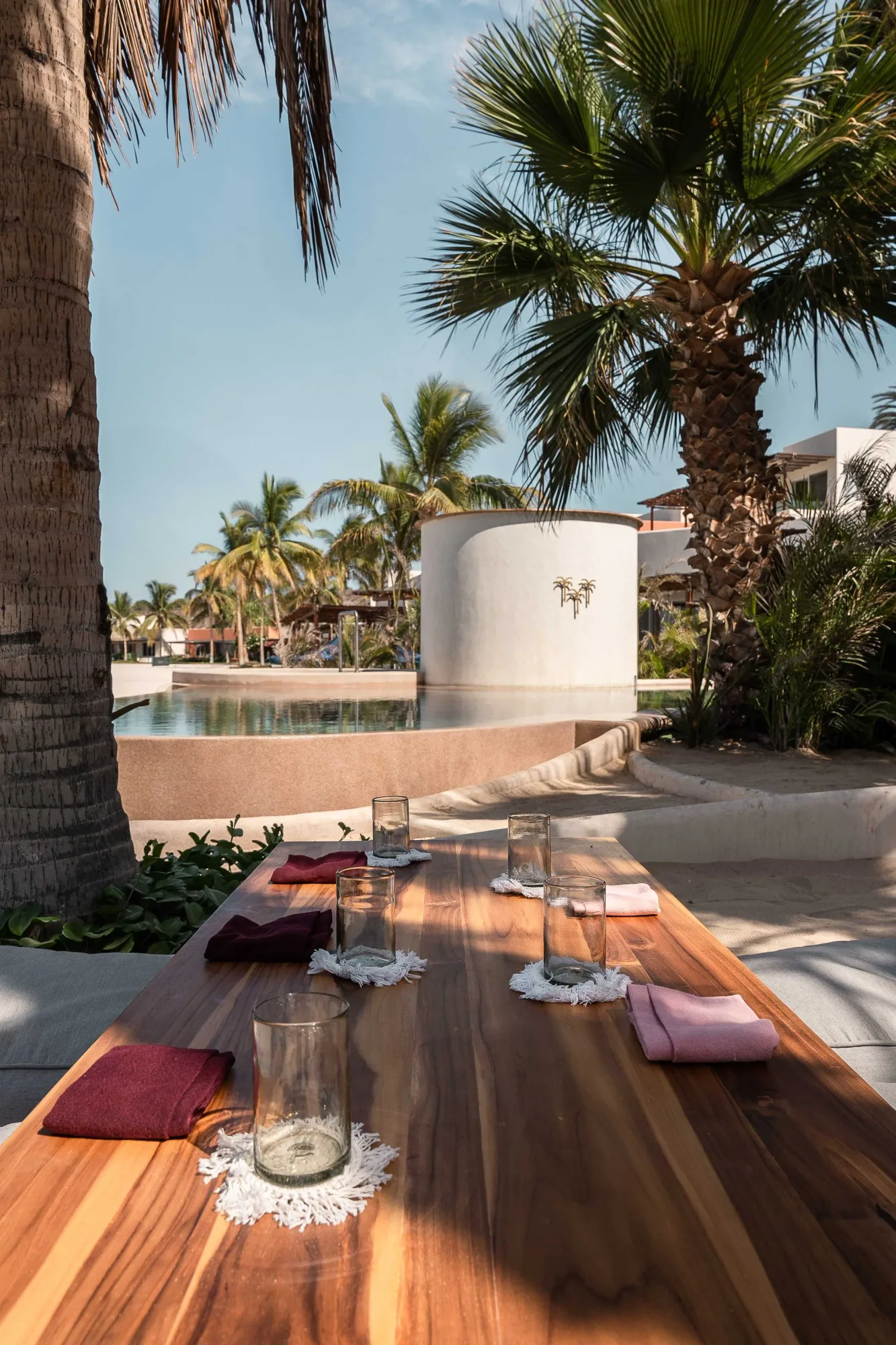 Long teak communal table set with fringed candle holders and printed napkins by the pool, palms and a round planter beyond