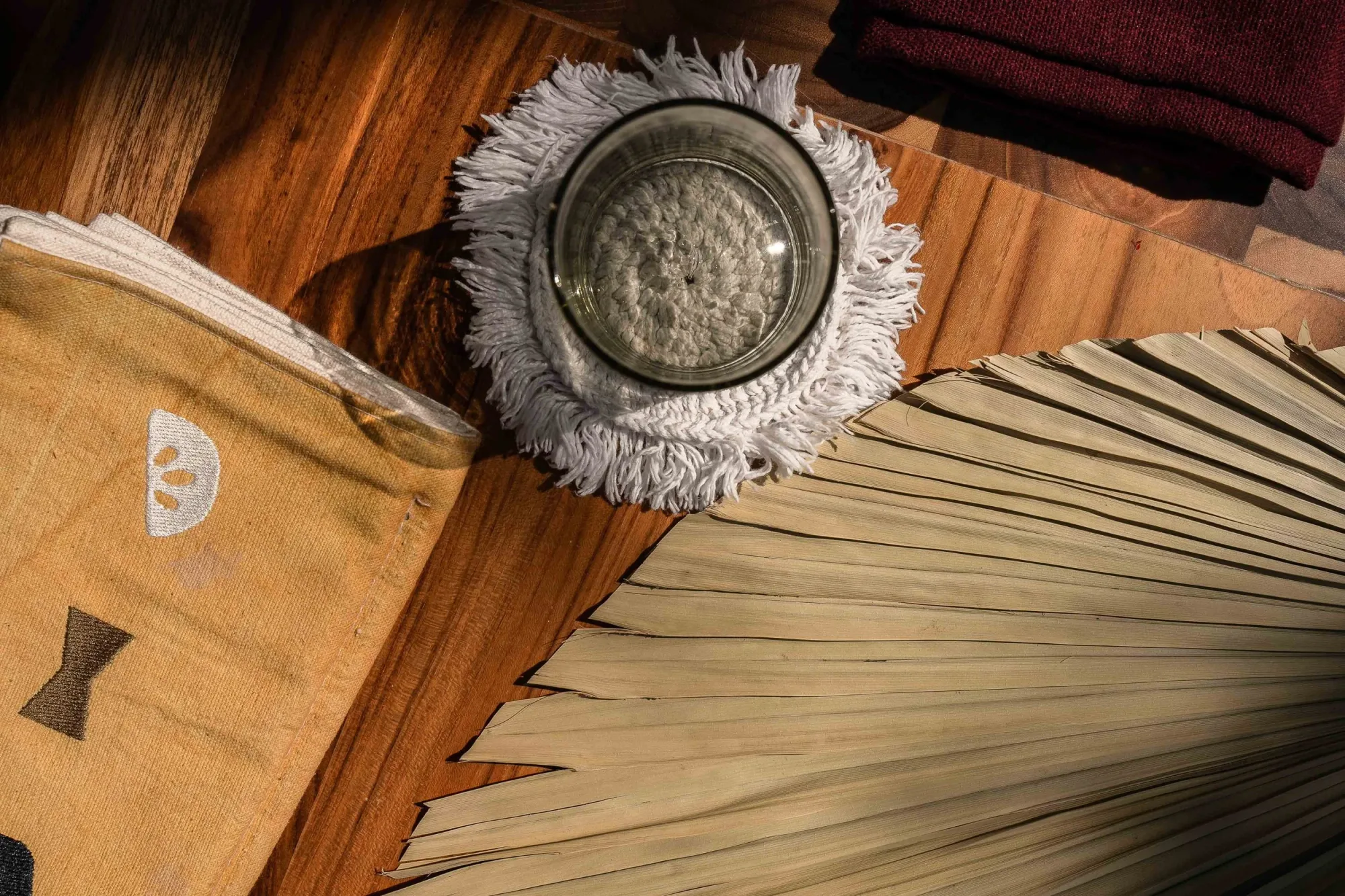 Overhead detail of a fringed glass candle holder on teak beside a dried palm fan and a printed napkin with the logo