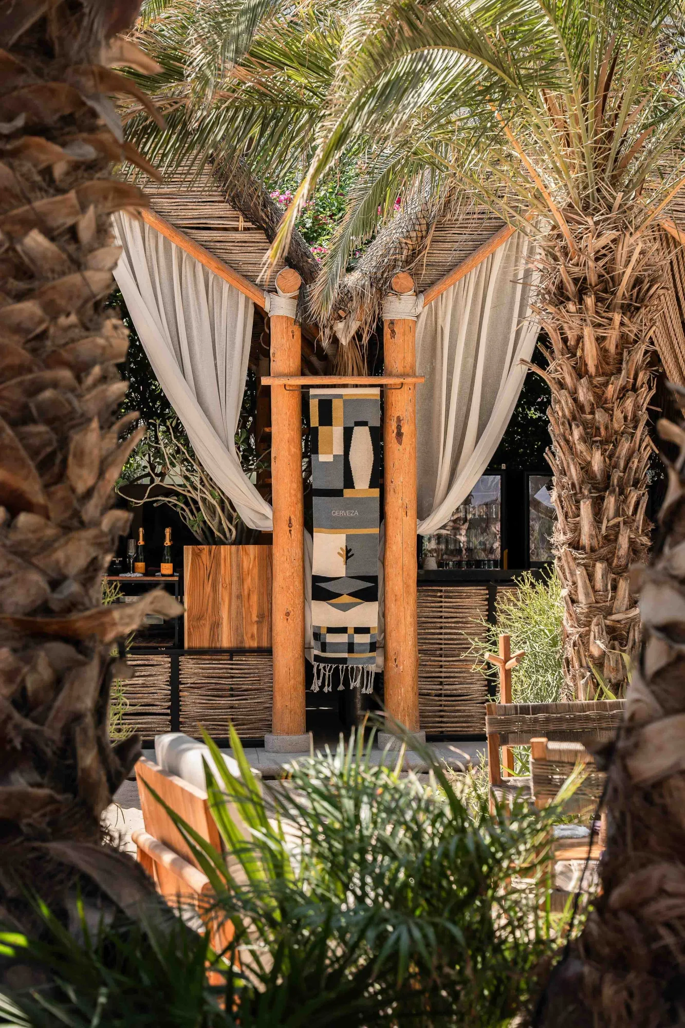 Palapa cabana framed between palms, with white linen curtains, timber posts, a graphic textile and low seating