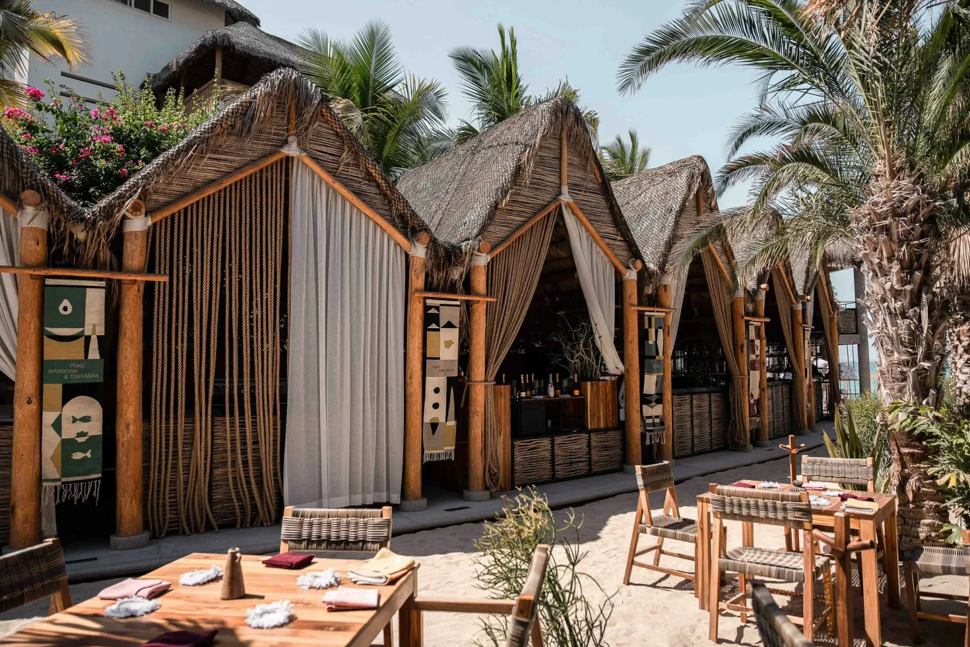 Row of palapa cabanas with thatched gables, rope and linen curtains and the bar, teak dining tables set on the sand
