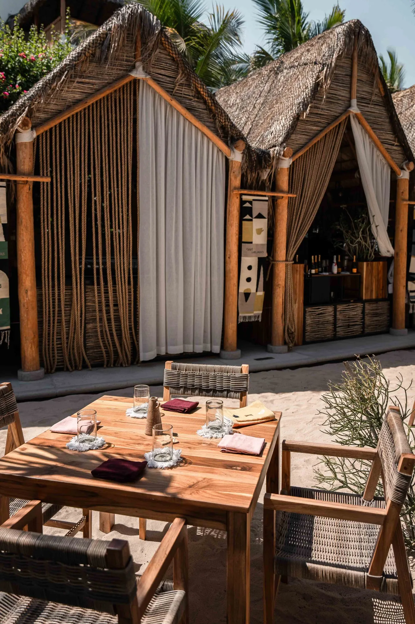 Palapa cabanas and the bar behind a set teak dining table with woven chairs on the sand