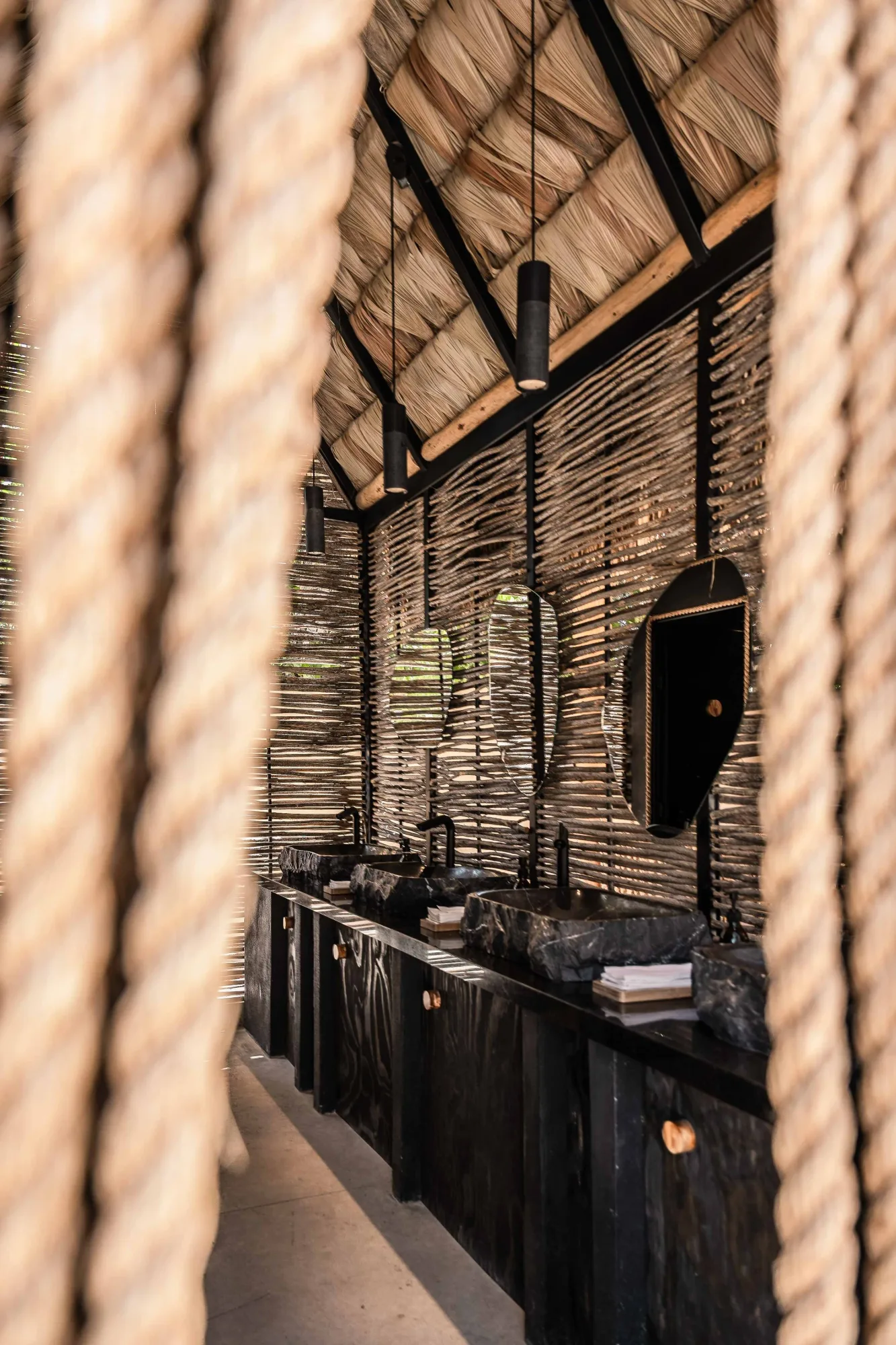 Restroom under a thatched roof with black ocotillo-stick walls, a dark vanity with stone vessel sinks and cone pendants